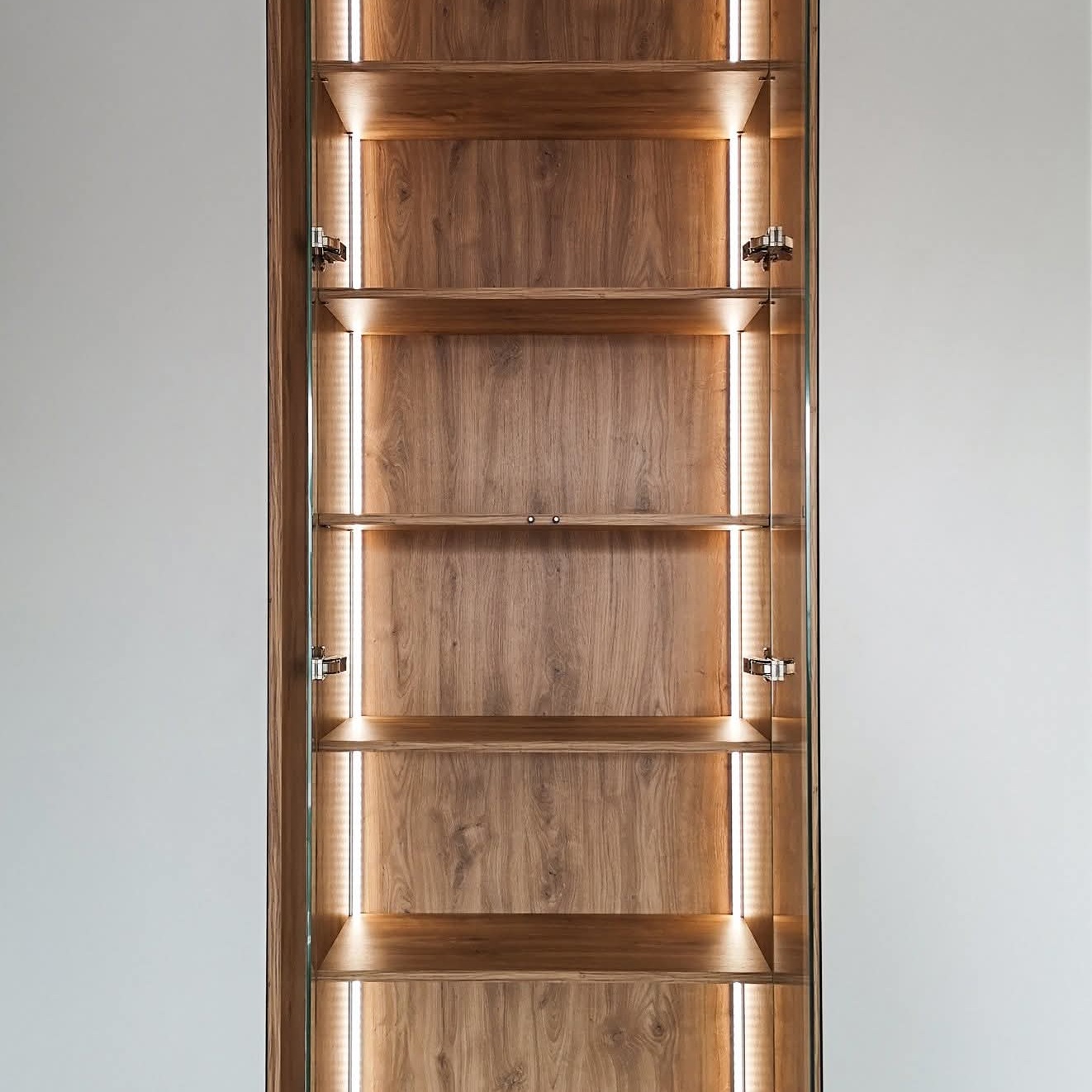 Otwarta szafa wnękowa z drewnianymi półkami i oświetleniem LED po bokach, zawiasy widoczne na szklanych drzwiach, minimalistyczny design.