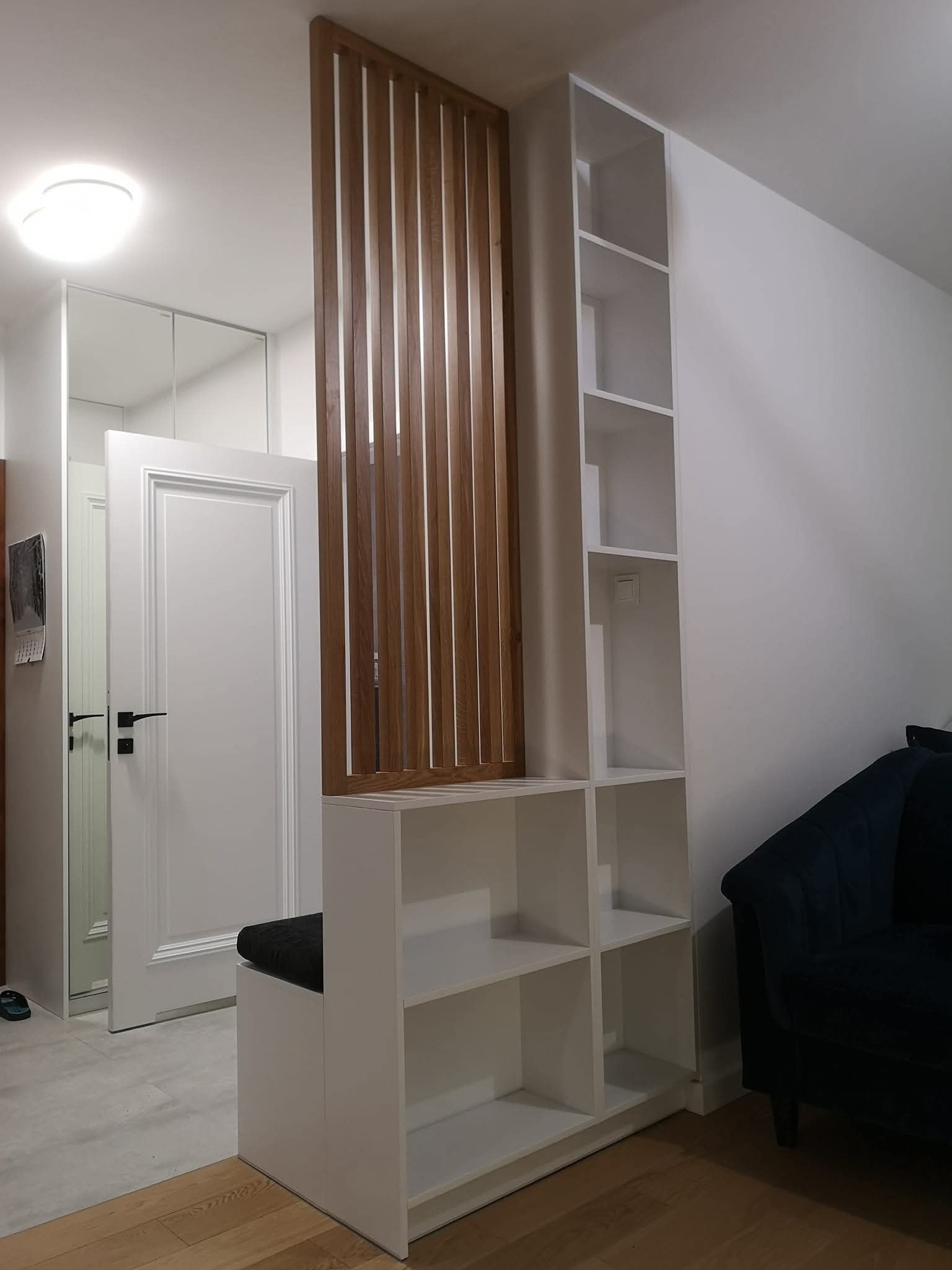 Nowoczesna zabudowa przedpokoju: białe szafki z drewnianą ażurową ścianką działową, siedzisko i wysoka półka. Minimalistyczny design, jasne wnętrze.