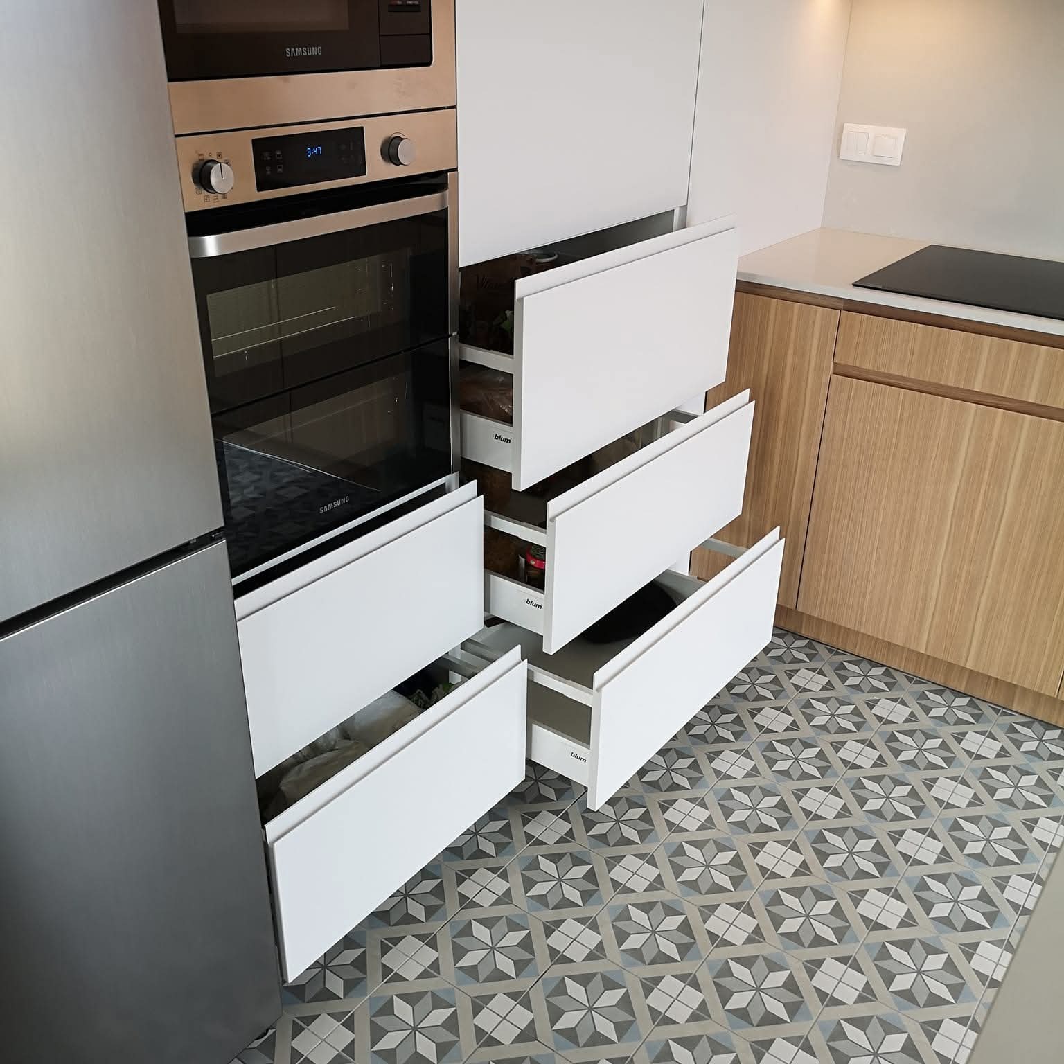 Nowoczesna kuchnia: zabudowa z piekarnikiem Samsung, białe szuflady Blum częściowo wysunięte, drewnopodobne szafki i wzorzysta podłoga. Ujęcie z bliska.