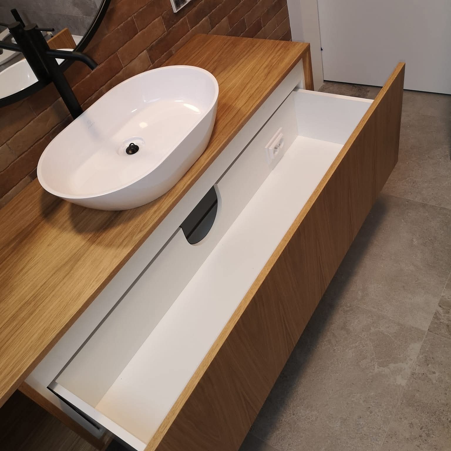 Nowoczesna szafka łazienkowa z owalną umywalką nablatową, drewnianym blatem i wysuwaną szufladą z gniazdkiem. Ceglana ściana i szara podłoga w tle.
