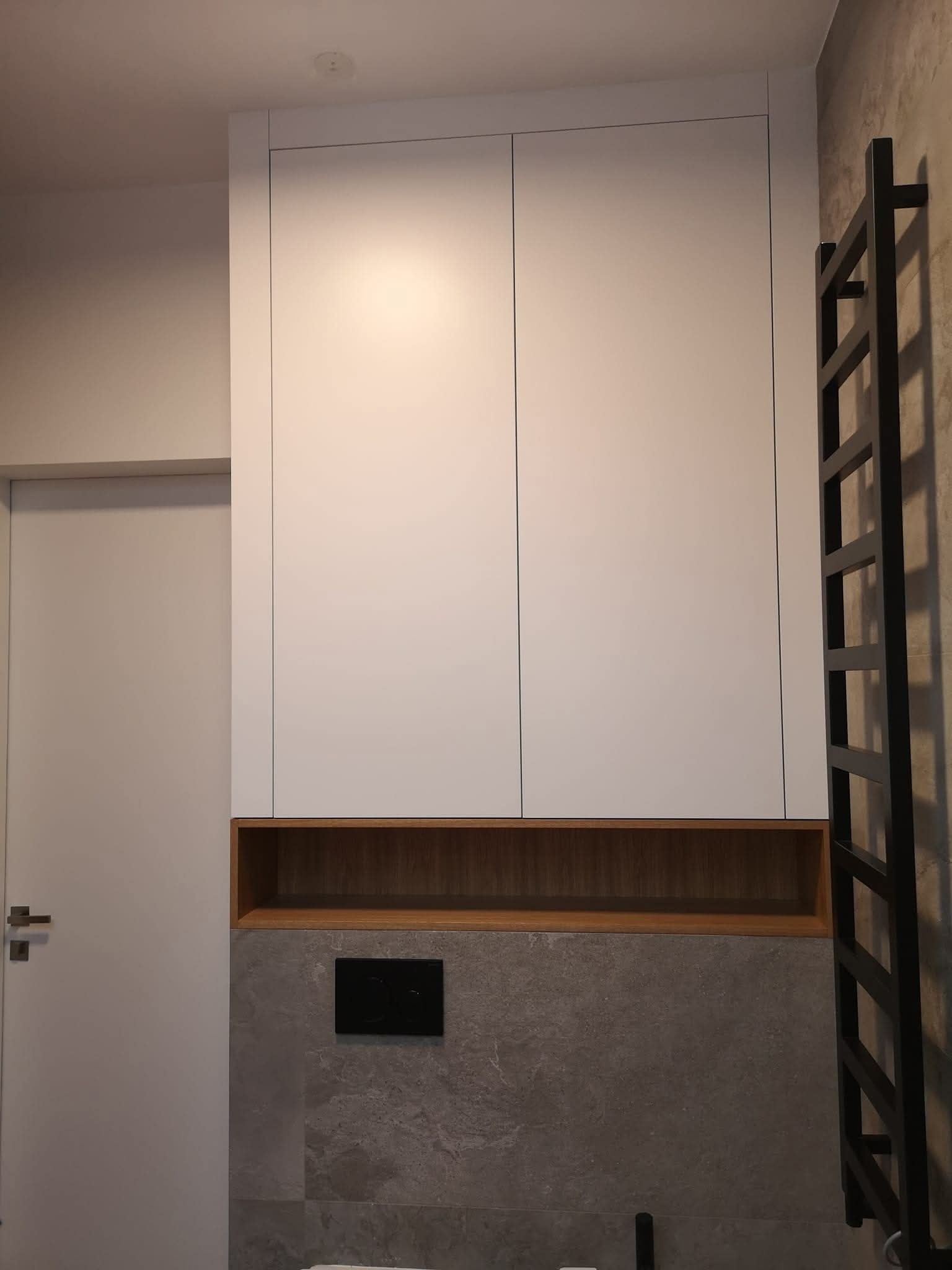 Biała szafka łazienkowa z drewnianą wnęką, szara ściana z czarnym przyciskiem spłuczki, grzejnik drabinkowy i fragment drzwi po lewej stronie.