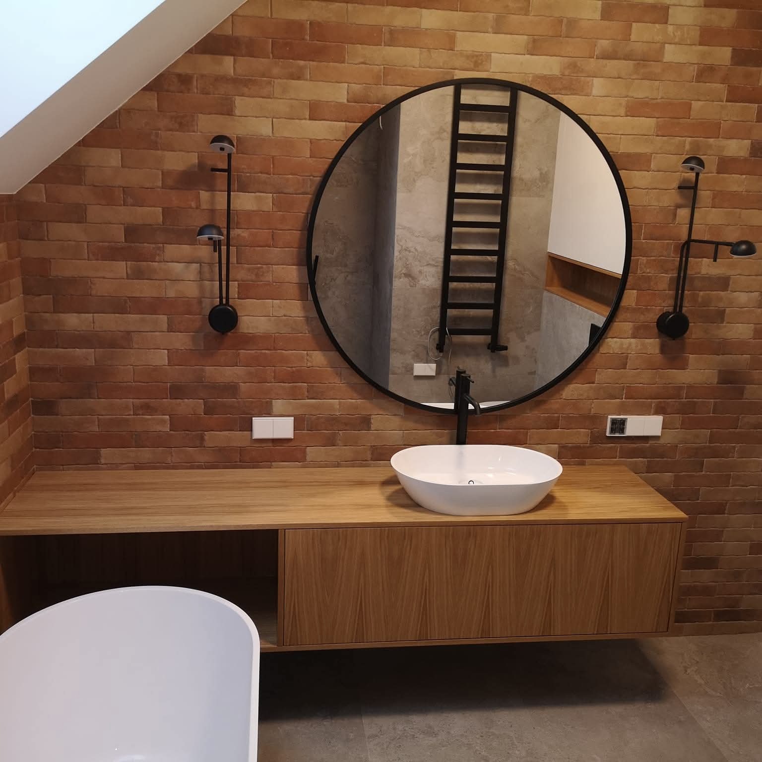 Nowoczesna łazienka z umywalką nablatową na drewnianej szafce, okrągłym lustrem i ceglaną ścianą. Minimalistyczne oświetlenie i grzejnik drabinkowy w tle.