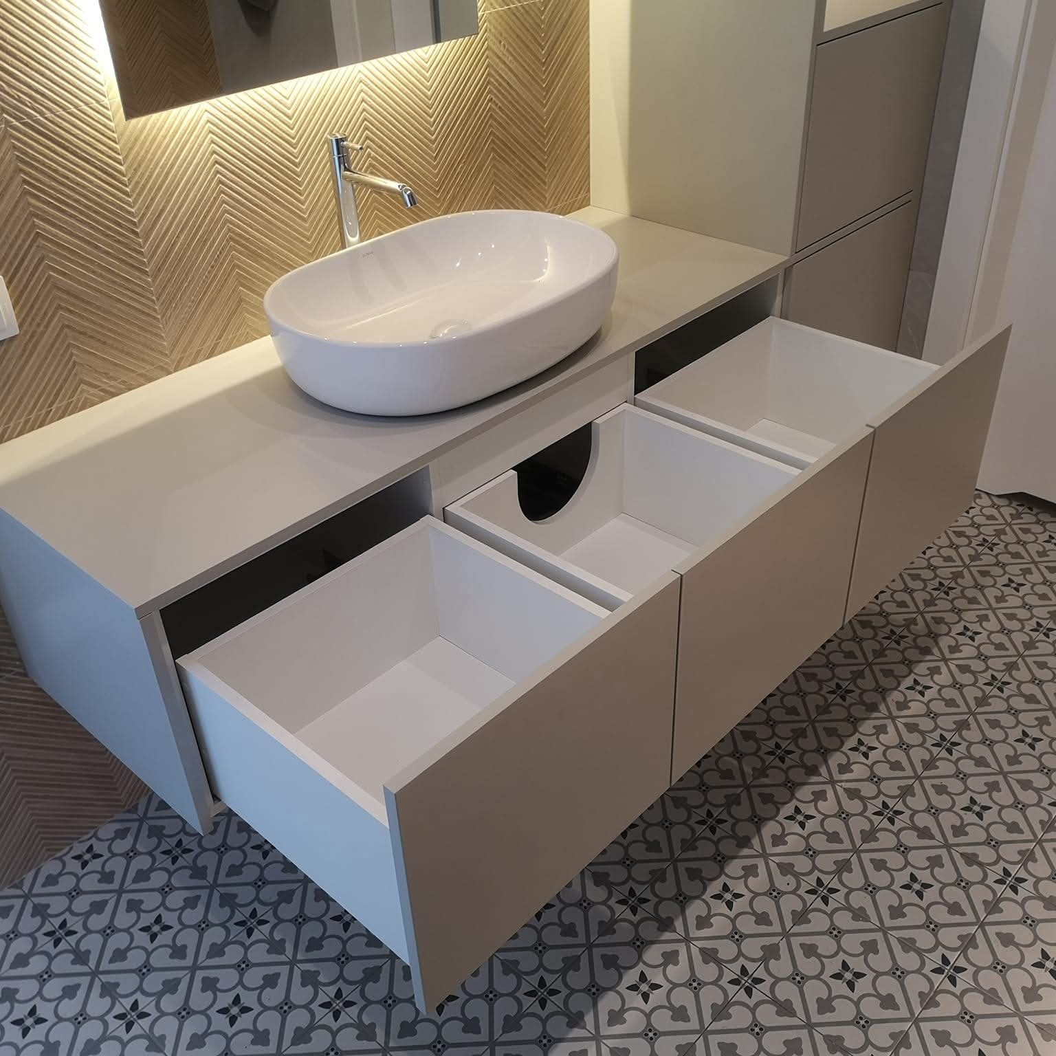 Biała, owalna umywalka nablatowa z chromowaną baterią na szarej szafce z trzema wysuwanymi szufladami. W tle dekoracyjna ściana i wzorzysta podłoga.