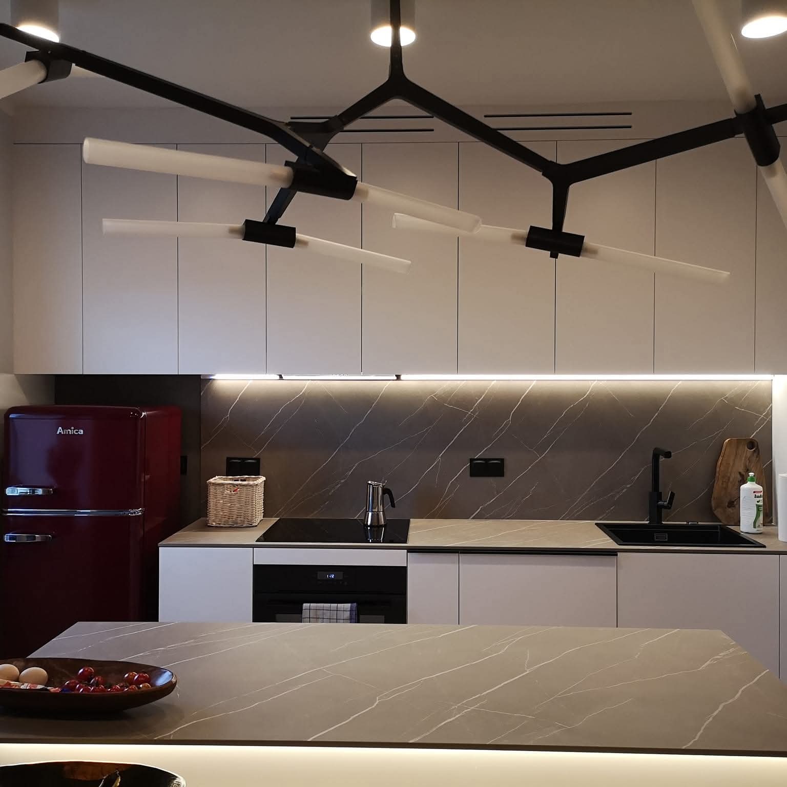 Nowoczesna kuchnia z wyspą, marmurowym blatem, retro lodówką Amica i awangardową lampą sufitową. Minimalistyczne szafki i subtelne oświetlenie LED.