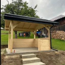 Nowa drewniana altana z czarnym dachem i rynną, betonowe schody wejściowe, na zewnątrz trawa i drzewa. W tle sterta drewna i osoby przy pracy.
