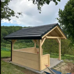 Drewniana altana z czarnym dachem na kostce brukowej, otoczona zielenią. W tle las i błękitne niebo. Wózek budowlany z łopatą w trawie. Perspektywa z dołu, widok na konstrukcję.