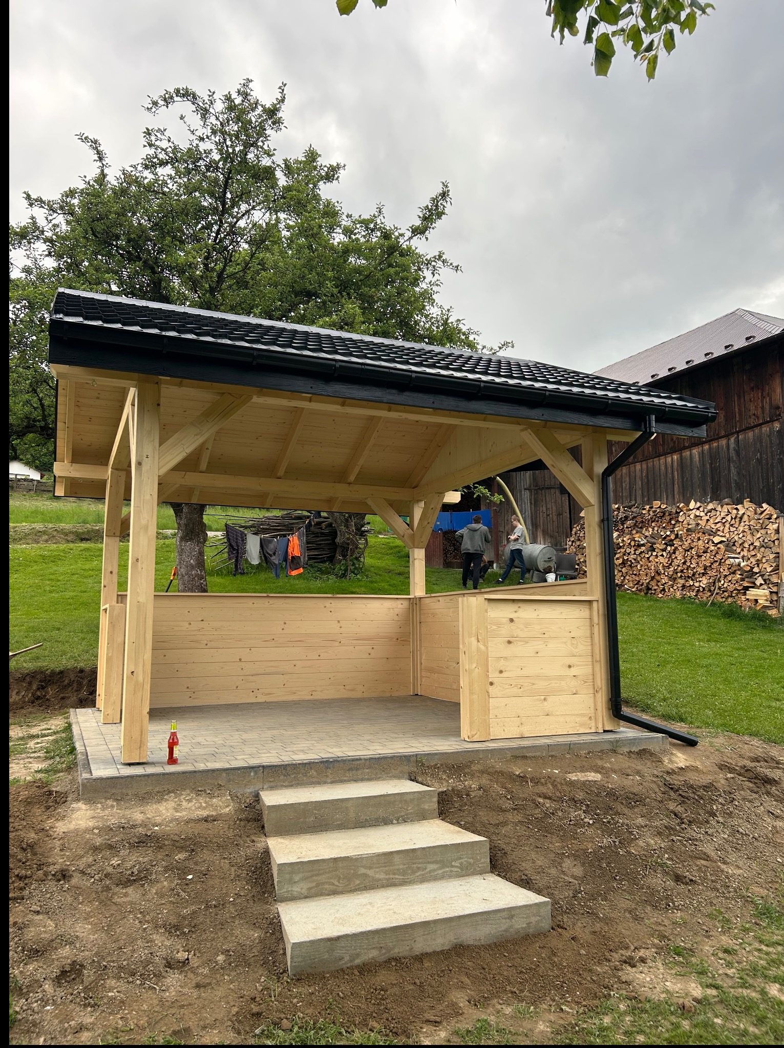 Nowa drewniana altana z czarnym dachem i rynną, betonowe schody wejściowe, na zewnątrz trawa i drzewa. W tle sterta drewna i osoby przy pracy.