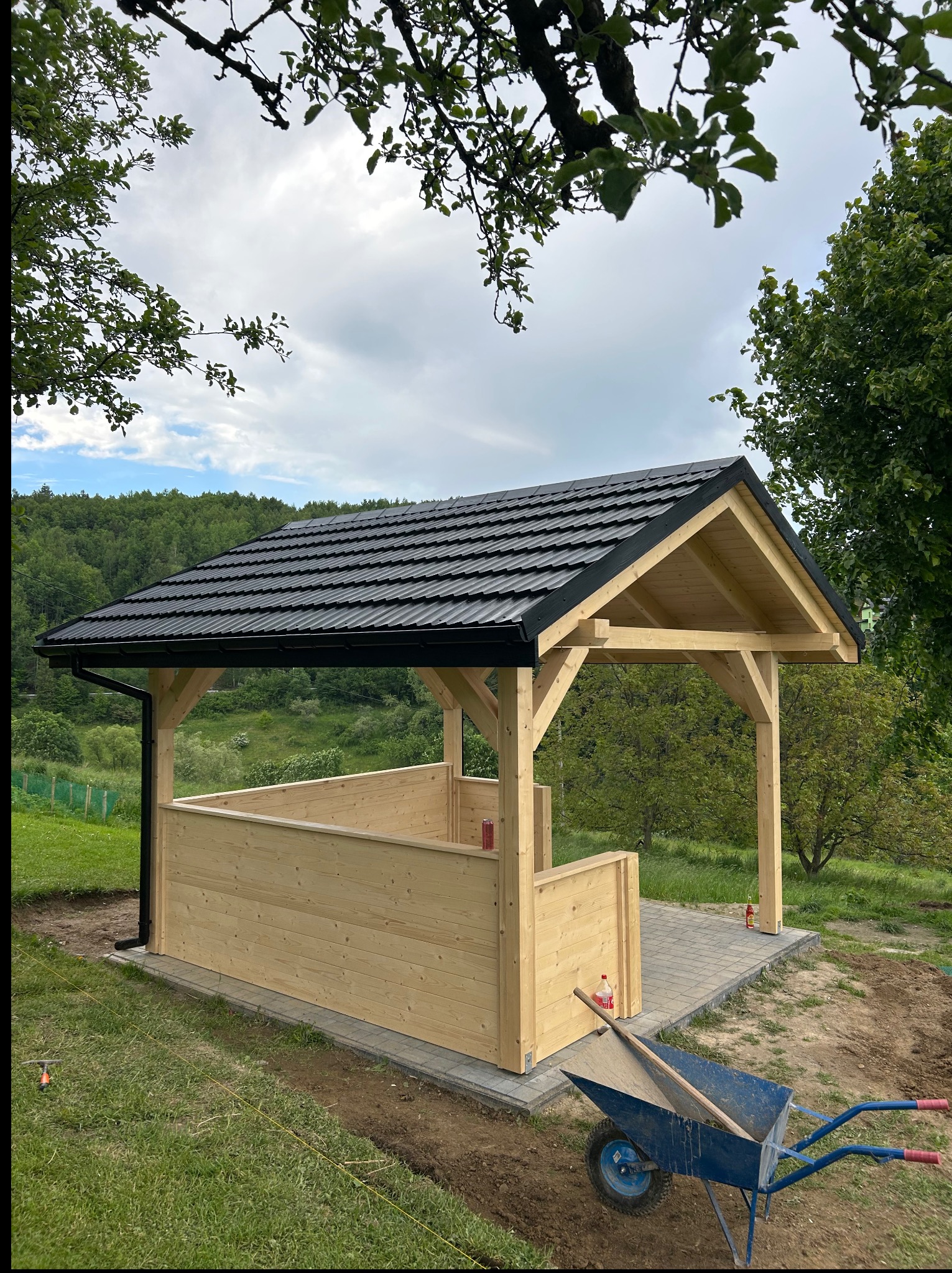 Drewniana altana z czarnym dachem na kostce brukowej, otoczona zielenią. W tle las i błękitne niebo. Wózek budowlany z łopatą w trawie. Perspektywa z dołu, widok na konstrukcję.