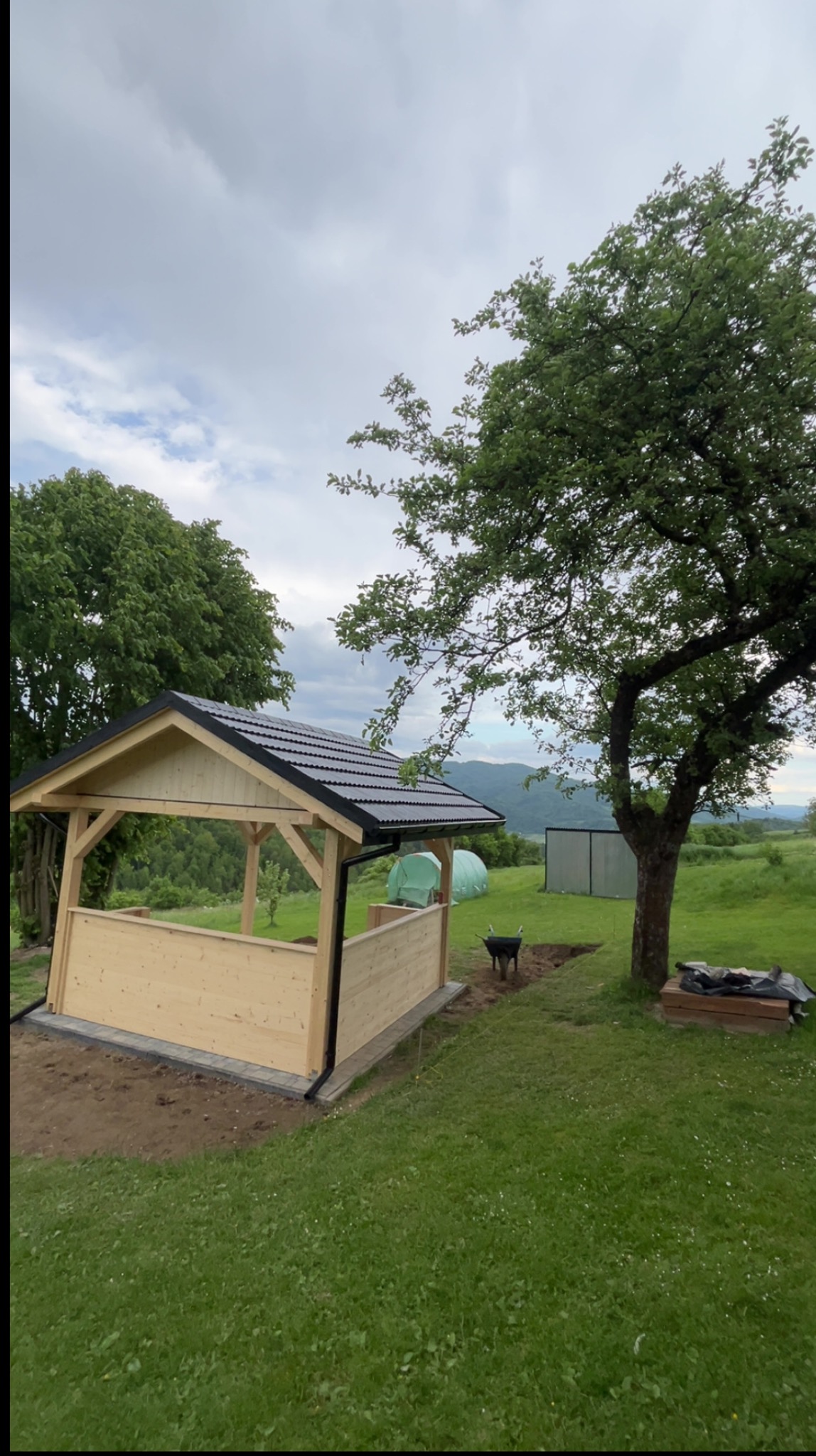 Drewniana altana z czarnym dachem na trawie, w tle góry i drzewa. Widoczna taczka i przygotowany teren. Kąt ujęcia z dołu podkreśla solidność konstrukcji.