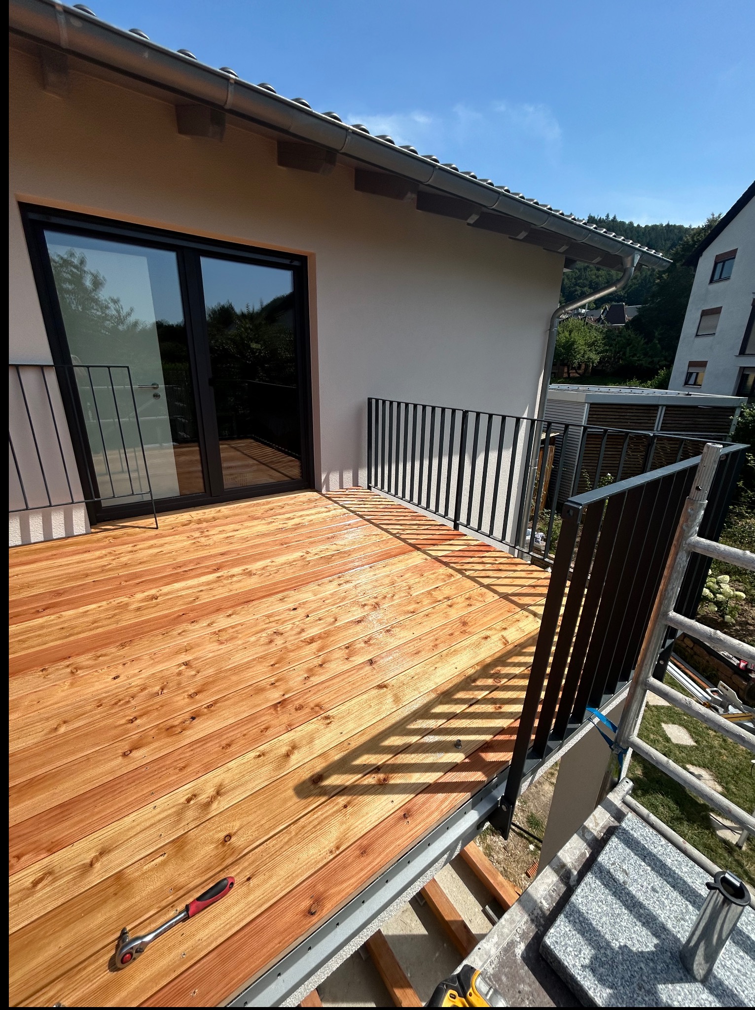 Nowy, drewniany taras z czarną balustradą przy budynku. Na tarasie leży klucz nasadowy. Widoczna drabina i elementy montażowe. Słoneczny dzień.