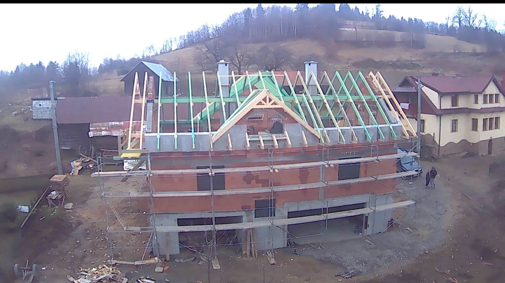 Budowa domu jednorodzinnego z rusztowaniem, widok z góry na konstrukcję dachu i ceglaną elewację w trakcie wznoszenia na tle pagórkowatego krajobrazu.