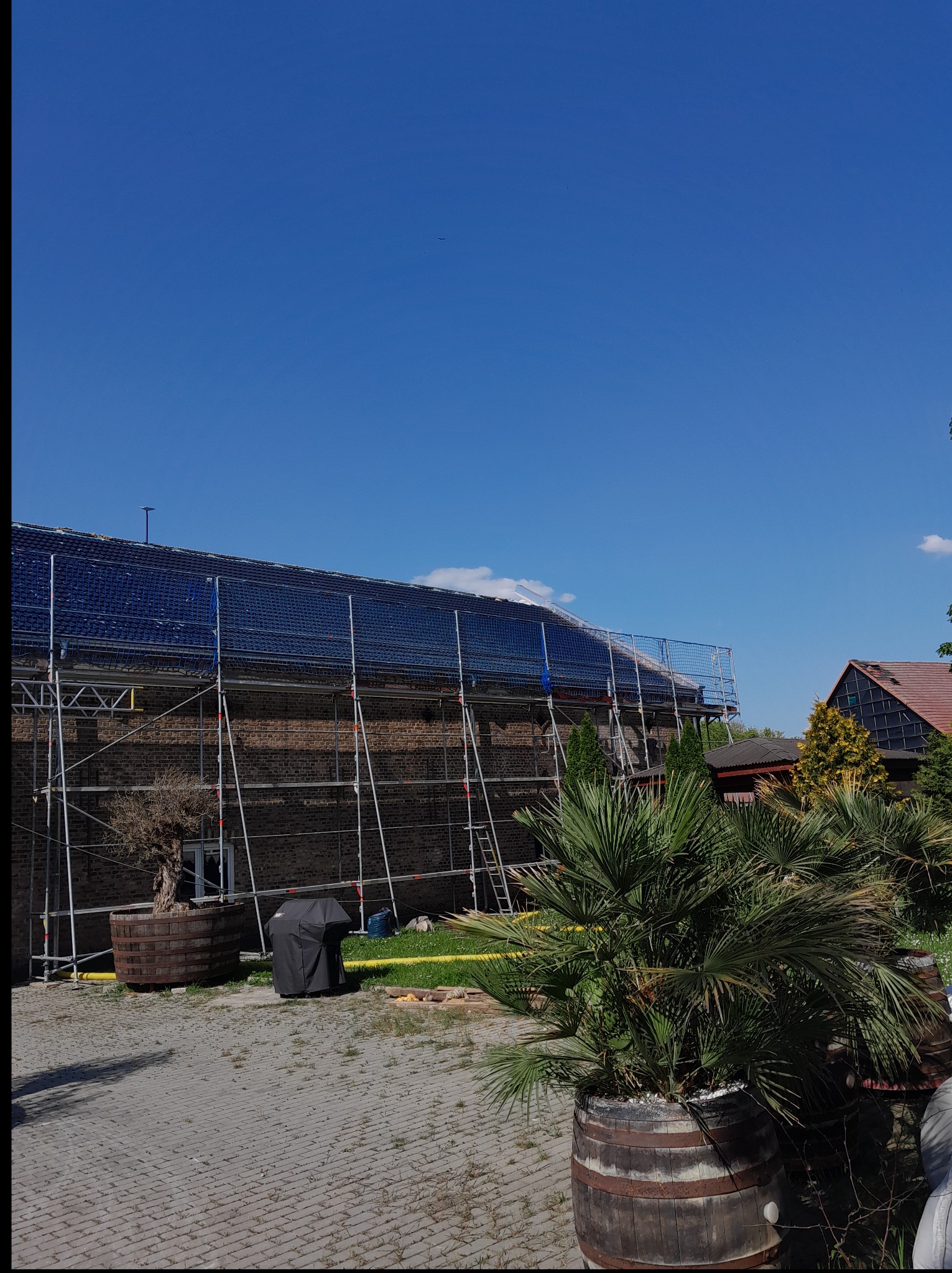 Dom z cegły otoczony rusztowaniami i siatką zabezpieczającą, widoczny fragment dachu. Na pierwszym planie roślinność w donicach, słoneczny dzień.