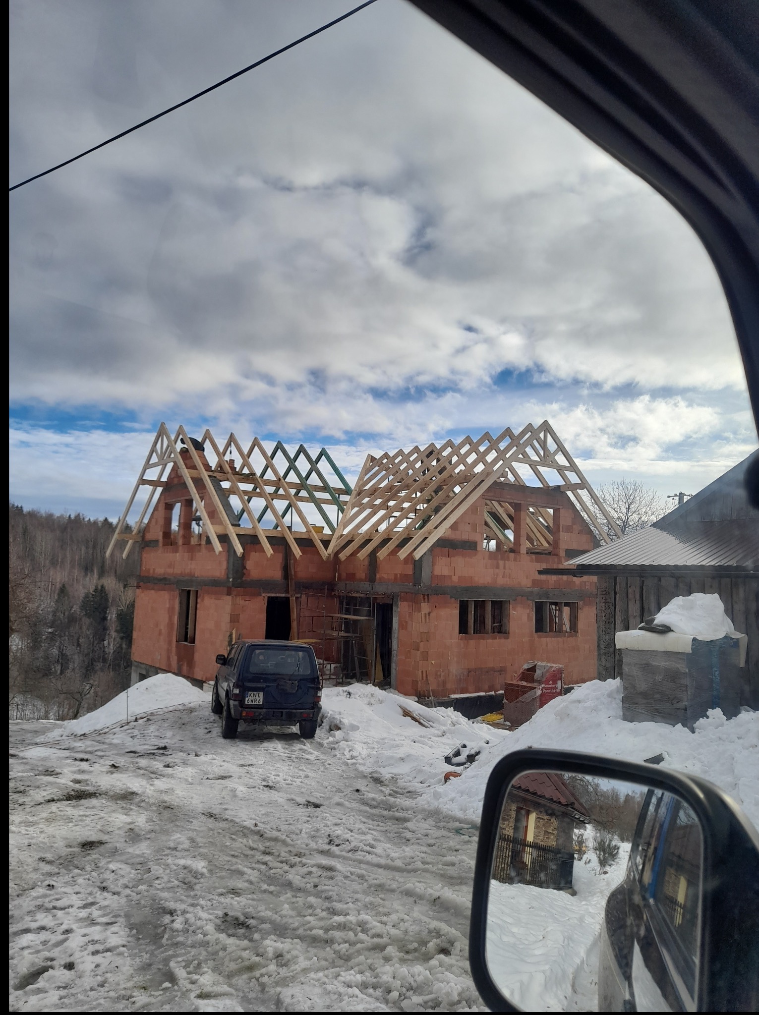 Surowy stan domu z czerwonej cegły z drewnianą konstrukcją dachu w zimowym krajobrazie, widziany z wnętrza samochodu. W tle las i pochmurne niebo.