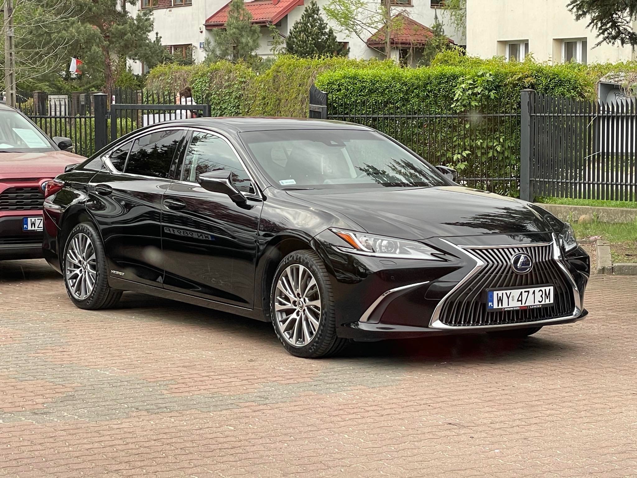 Czarna limuzyna Lexus ES na brukowanym podjeździe, z widoczną tablicą rejestracyjną i elementami chromowanymi. W tle budynki mieszkalne i zielony żywopłot.