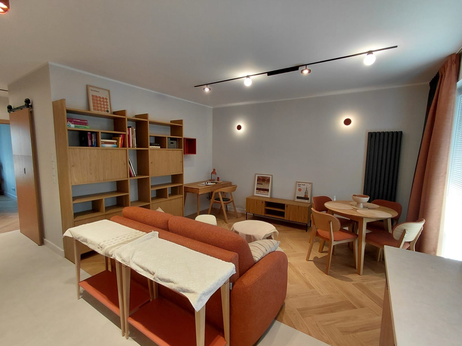 Jasne wnętrze salonu z parkietem w jodełkę, pomarańczową sofą i drewnianymi meblami. Widoczne oświetlenie punktowe i dekoracje ścienne.