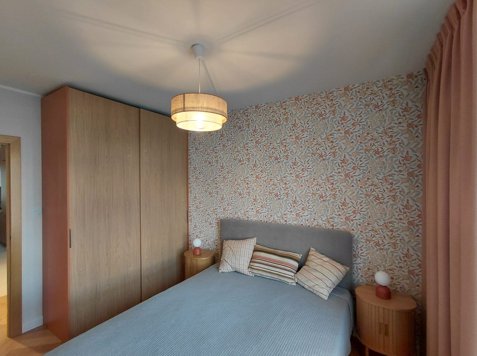 Ustawna sypialnia po remoncie: wzorzysta tapeta w odcieniach beżu i pomarańczy, drewniana szafa wnękowa, łóżko z szarym zagłówkiem i lampka nocna na szafce.