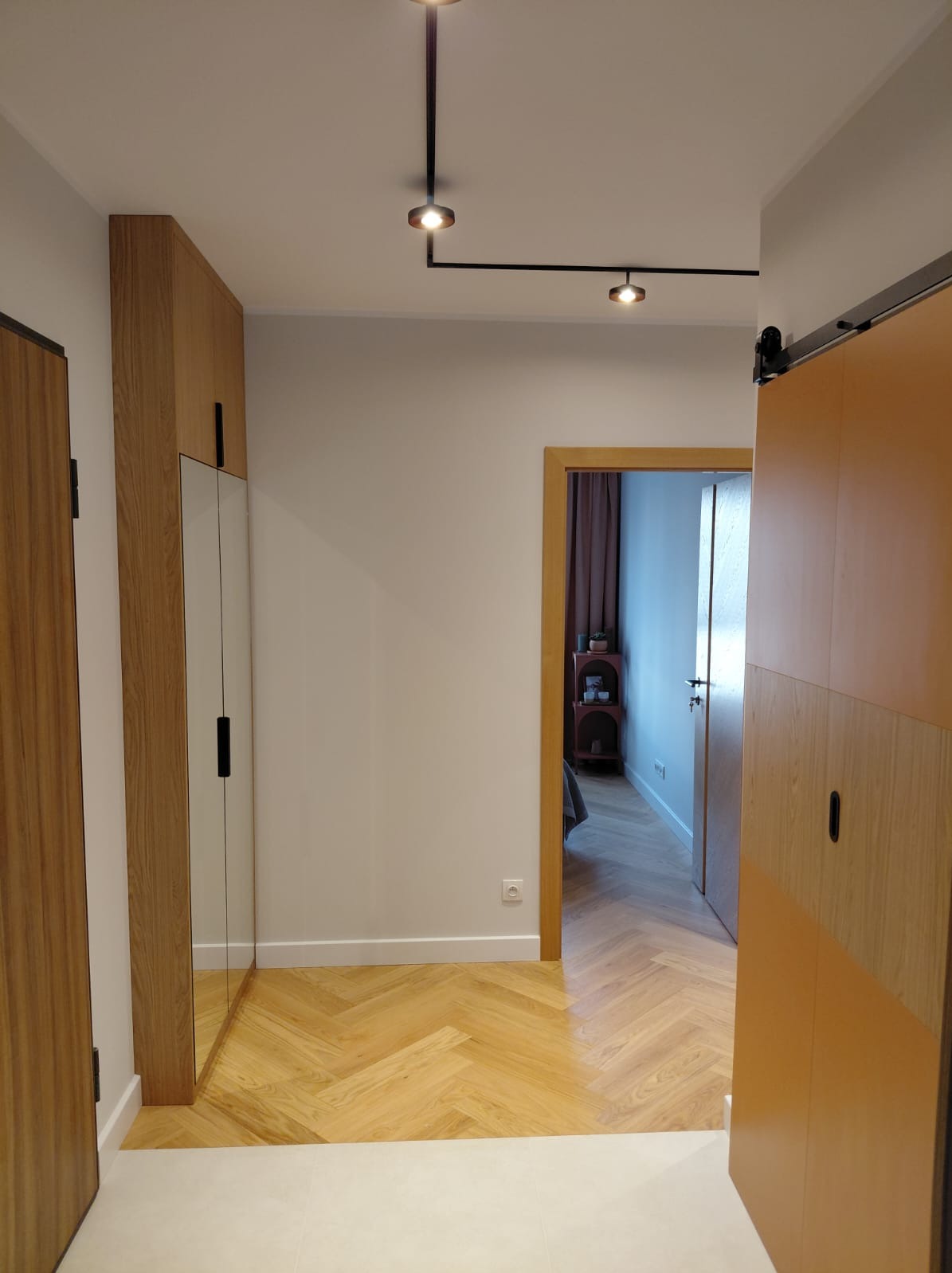 Nowoczesny przedpokój z parkietem w jodełkę, szafą z lustrem i przesuwnymi drzwiami. Widoczne oświetlenie punktowe i wejście do pokoju z elementami wyposażenia.
