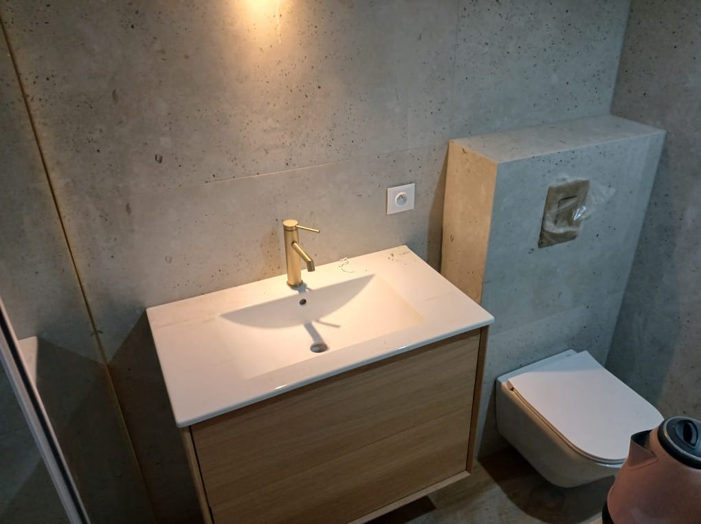 Nowoczesna łazienka z umywalką nablatową, złotą armaturą i płytkami imitującymi beton. Minimalistyczny design, wisząca toaleta i subtelne oświetlenie.