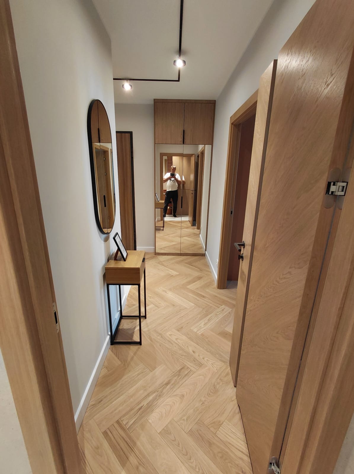 Jasny parkiet w jodełkę w korytarzu z lustrzaną szafą i owalnym lustrem. Widoczne drzwi z ukrytymi zawiasami i minimalistyczne oświetlenie sufitowe.