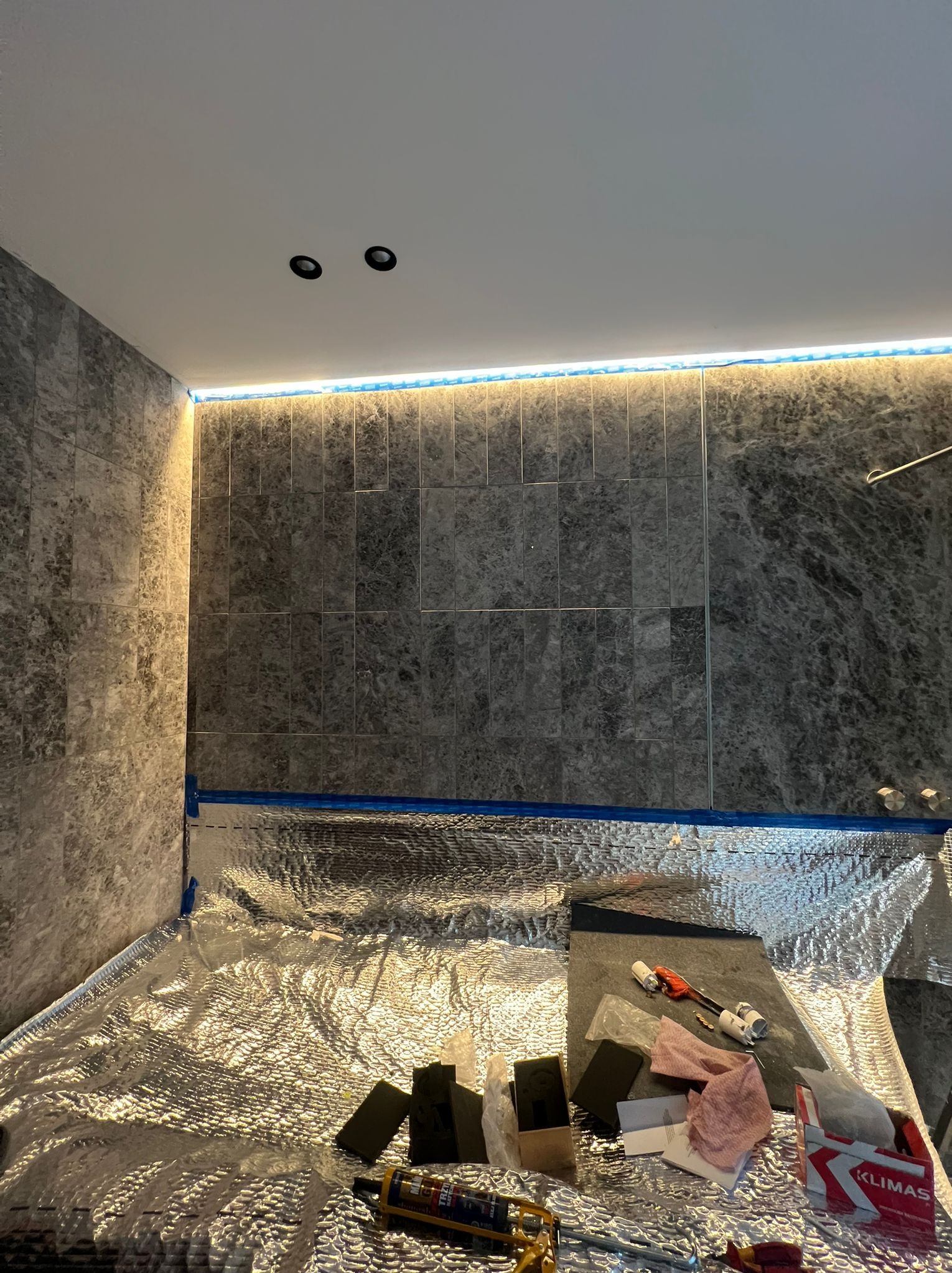 Łazienka w trakcie wykończenia: szare płytki na ścianach, folia izolacyjna na podłodze, narzędzia i materiały budowlane. Widoczne oświetlenie LED. Etap prac wykończeniowych.