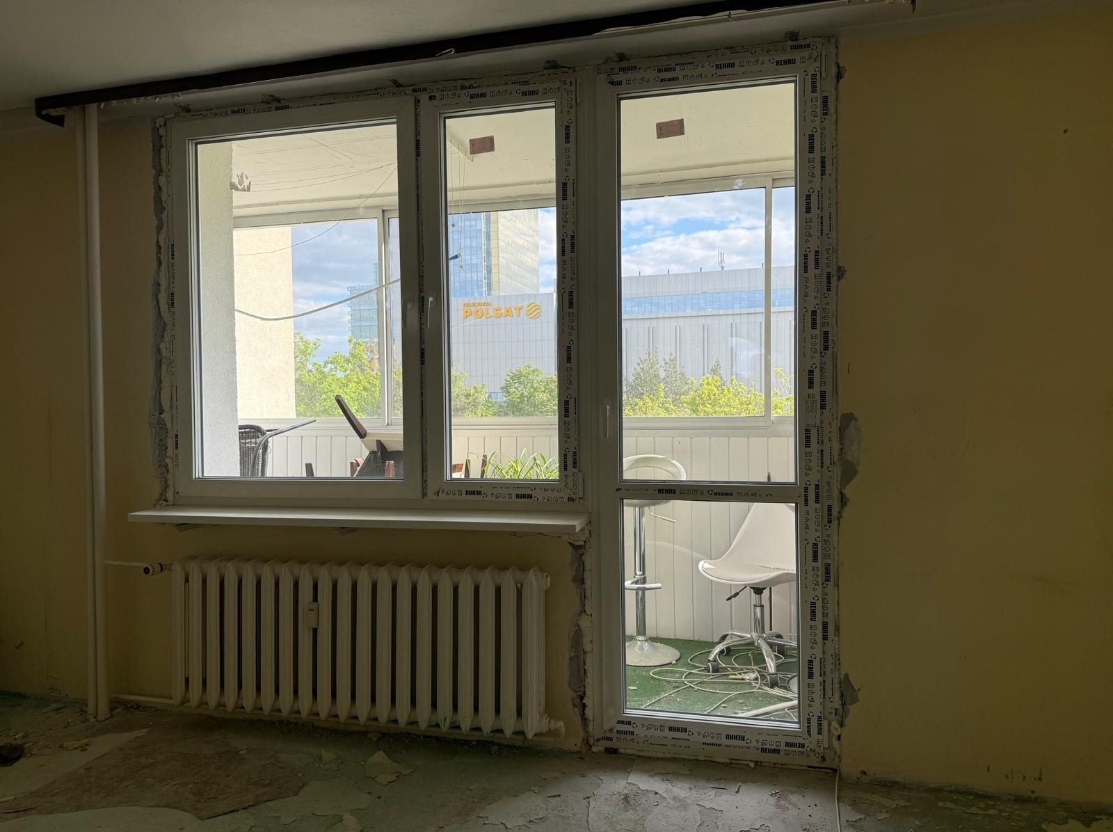 Wymiana okna balkonowego w mieszkaniu. Widoczne nowe okno PCV z widokiem na balkon i budynek Polsatu. Ściany w trakcie przygotowania do malowania, grzejnik. Etap prac wykończeniowych.
