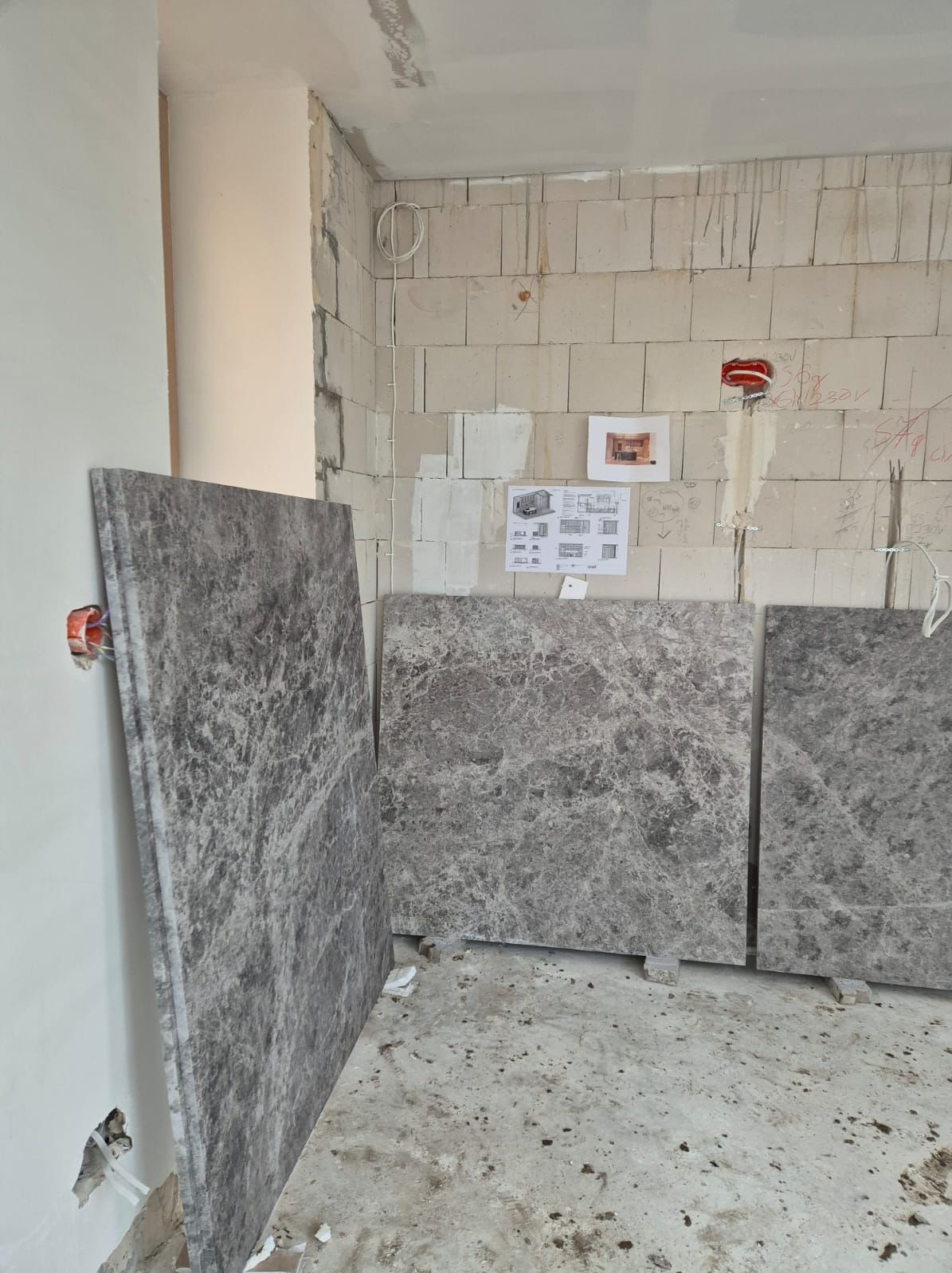 Płyty z szarego marmuru ustawione w surowym wnętrzu, przygotowane do montażu na ścianie z betonu komórkowego. Widoczne gniazdka elektryczne i plan pomieszczenia.
