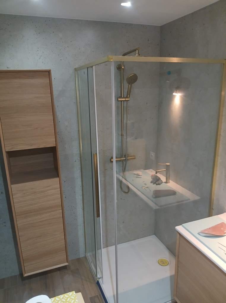Nowoczesna kabina prysznicowa z złotymi profilami i deszczownicą, obok szafka łazienkowa z jasnego drewna na tle ściany w odcieniach szarości. Wykończenie łazienki w minimalistycznym stylu.
