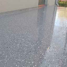 Artem Karpenko - Błyszcząca, szara posadzka żywiczna z białymi i czarnymi drobinkami, świeżo wylana na zewnątrz budynku. Widoczne rusztowania i ściana budynku.