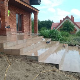 Artem Karpenko - Wyłożone jasnymi płytkami schody zewnętrzne domu jednorodzinnego, częściowo w ziemi. Mężczyzna ciągnie myjkę ciśnieniową. Widoczne narzędzia glazurnicze.