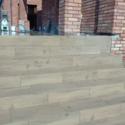 Kafelkowane schody wejściowe do domu, wykończone w odcieniach beżu z delikatnym wzorem drewna. W tle ceglana elewacja i myjka ciśnieniowa.