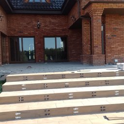 Artem Karpenko - Kaskadowe schody z płytek imitujących drewno, z widocznymi krzyżykami dystansowymi, prowadzące do wejścia domu z czerwonej cegły, w trakcie prac wykończeniowych.