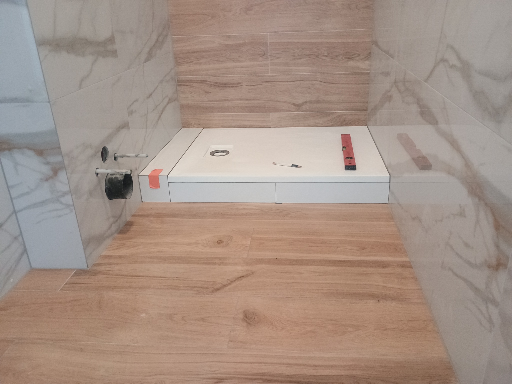 Biały brodzik z poziomnicą w trakcie montażu w łazience wykończonej płytkami imitującymi drewno i marmur. Widoczne przyłącza kanalizacyjne.