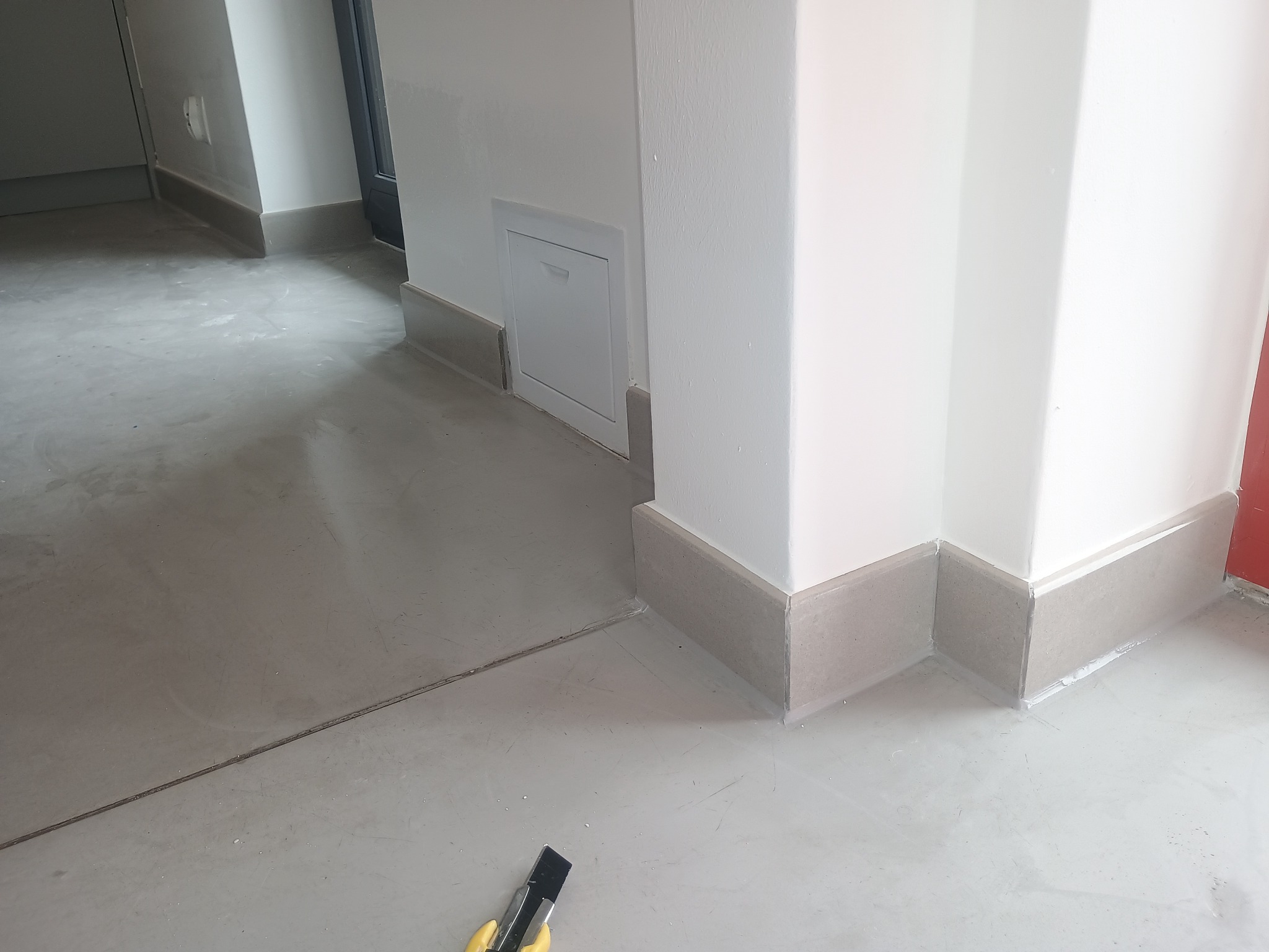 Wykończone szare listwy przypodłogowe w rogu pomieszczenia z szarą podłogą. Na podłodze leży nóż do tapet. Widoczna biała ściana i mała klapka rewizyjna.