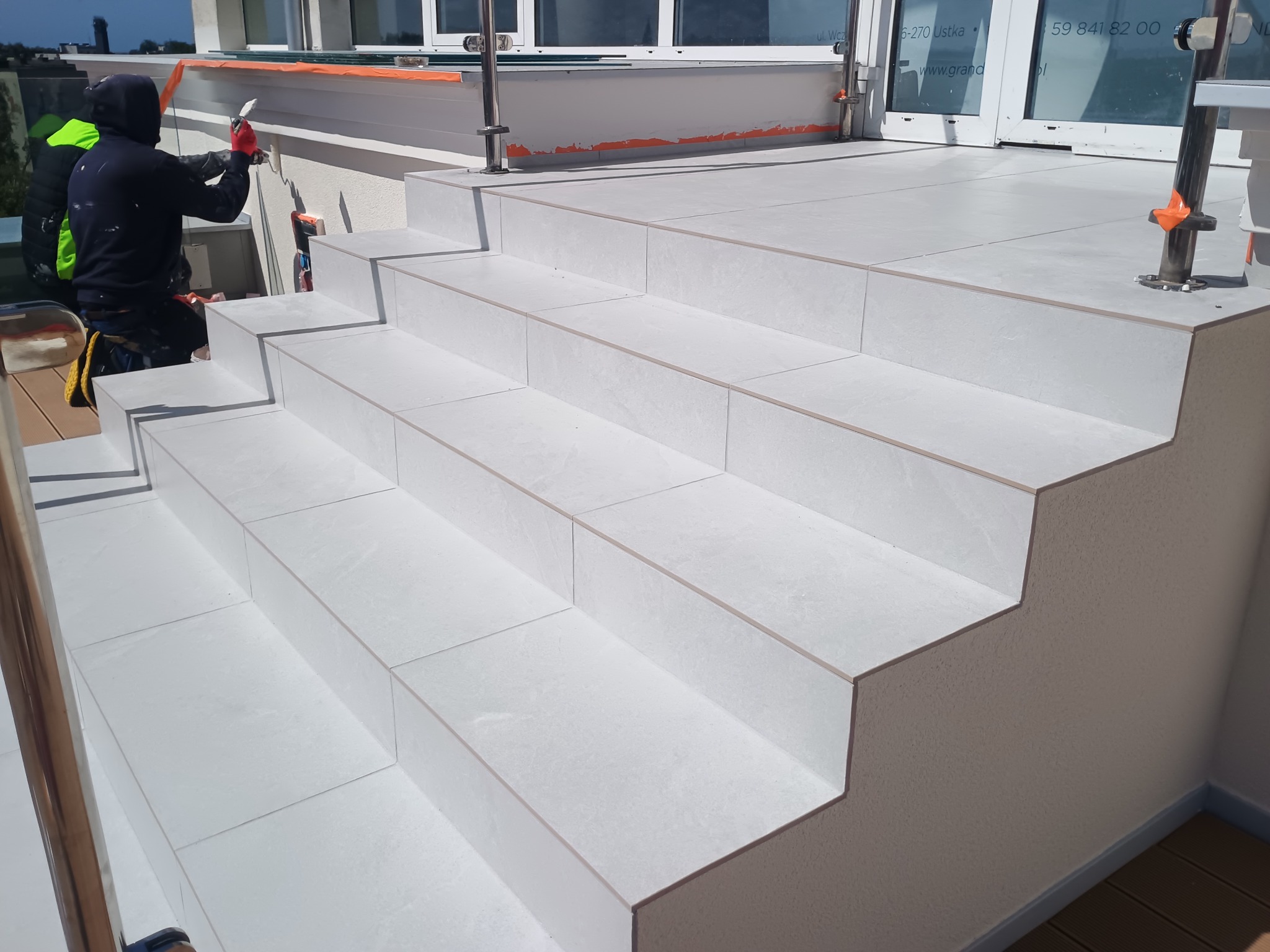 Białe płytki na schodach zewnętrznych, układane przez glazurnika w trakcie prac wykończeniowych. Widoczny pracownik w trakcie spoinowania płytek.