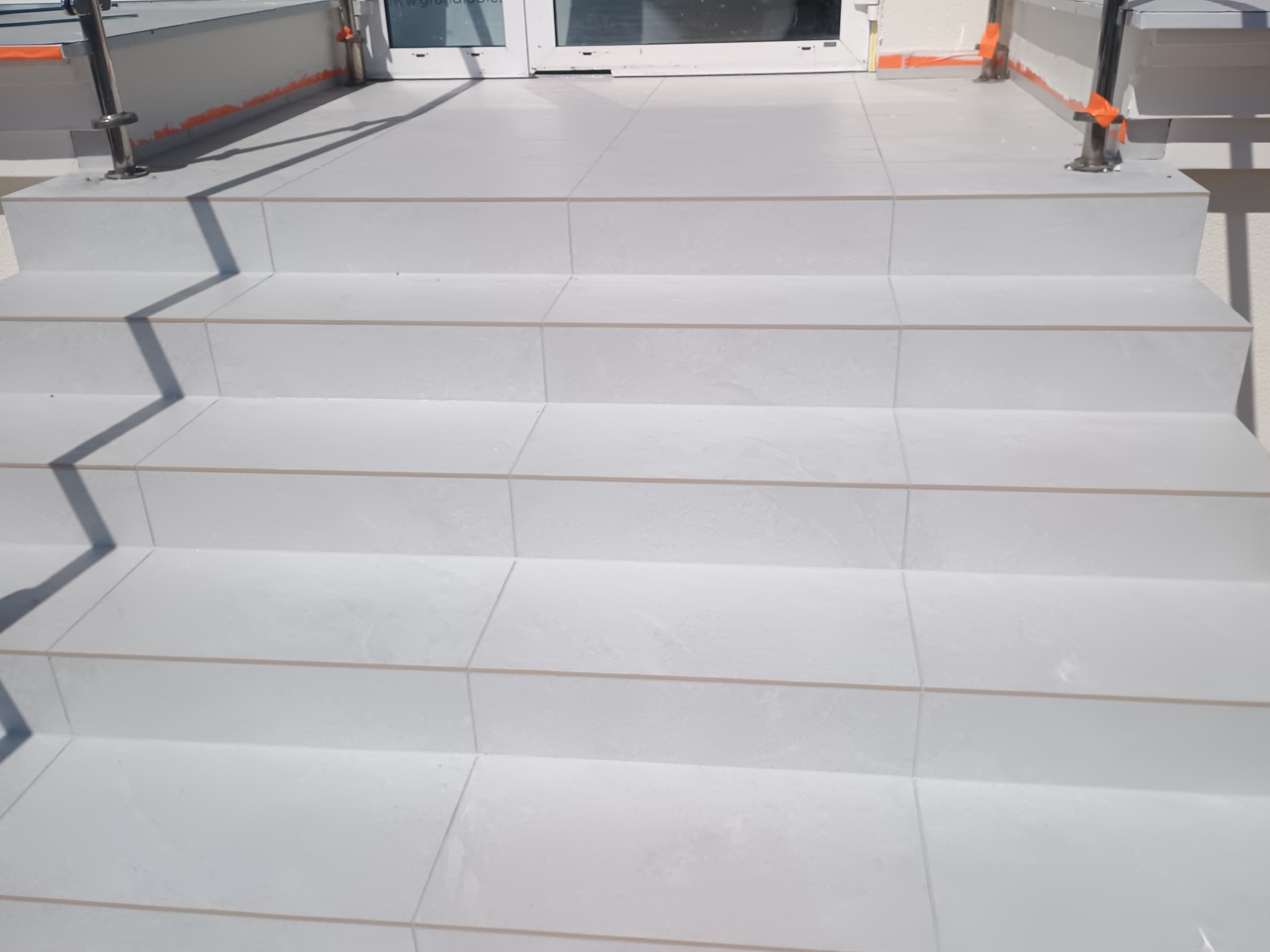 Białe płytki na schodach zewnętrznych, z widoczną fugą i fragmentem metalowej barierki. Minimalistyczny design, jasne oświetlenie.