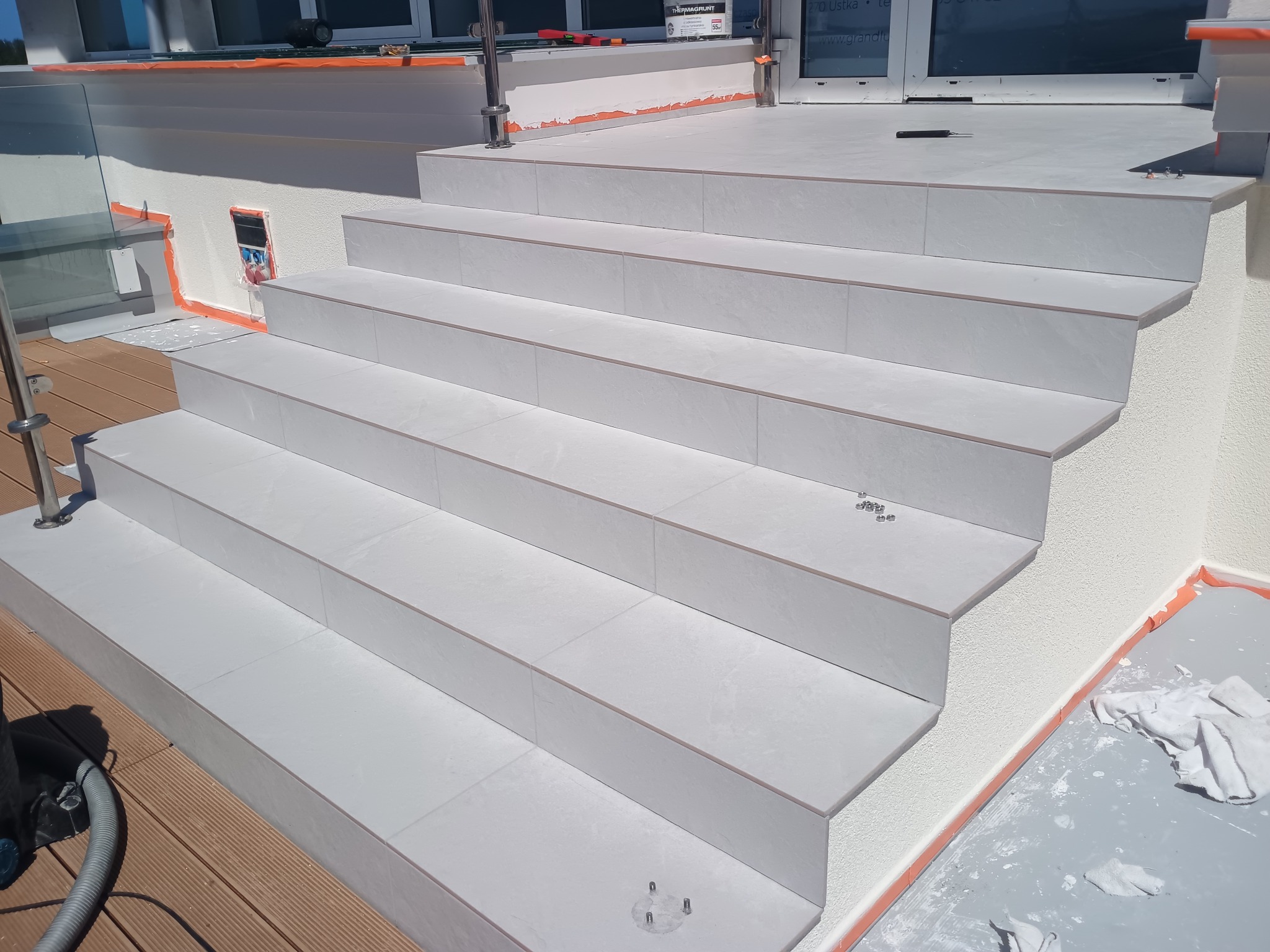 Wykończone schody zewnętrzne z jasnych płytek, z widocznymi detalami wykończeniowymi i elementami zabezpieczającymi. Widoczny fragment tarasu z deskami.