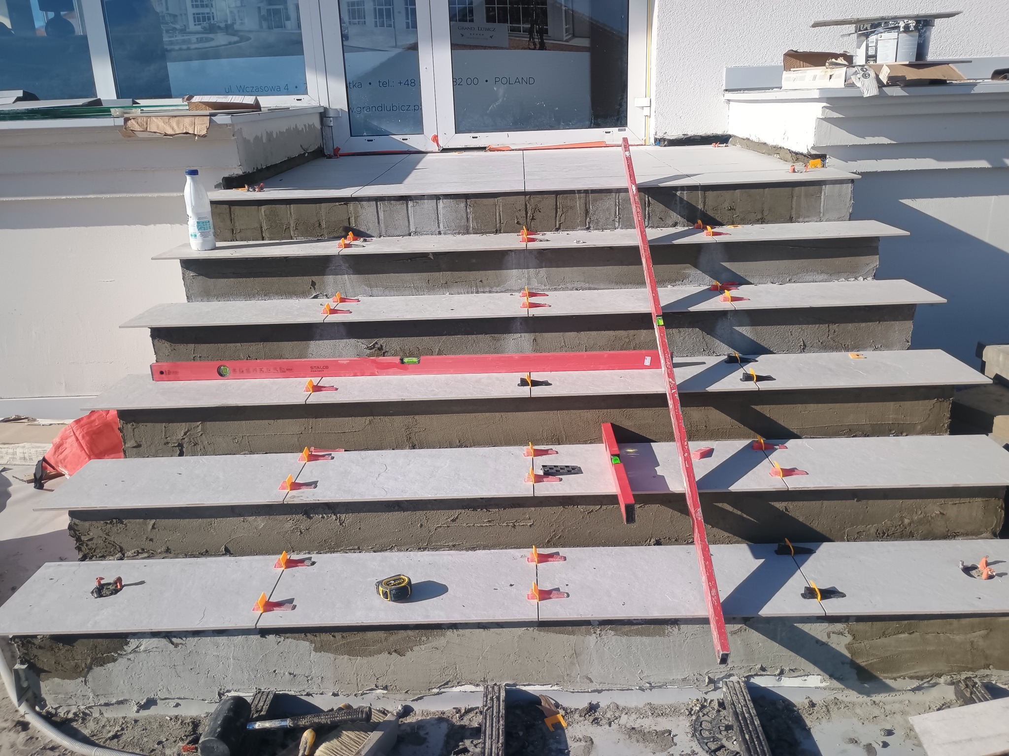 Kładzenie szarych płytek na schodach wejściowych, widoczne poziomice, krzyżyki dystansowe, klej. Prace glazurnicze w toku, budynek w tle.