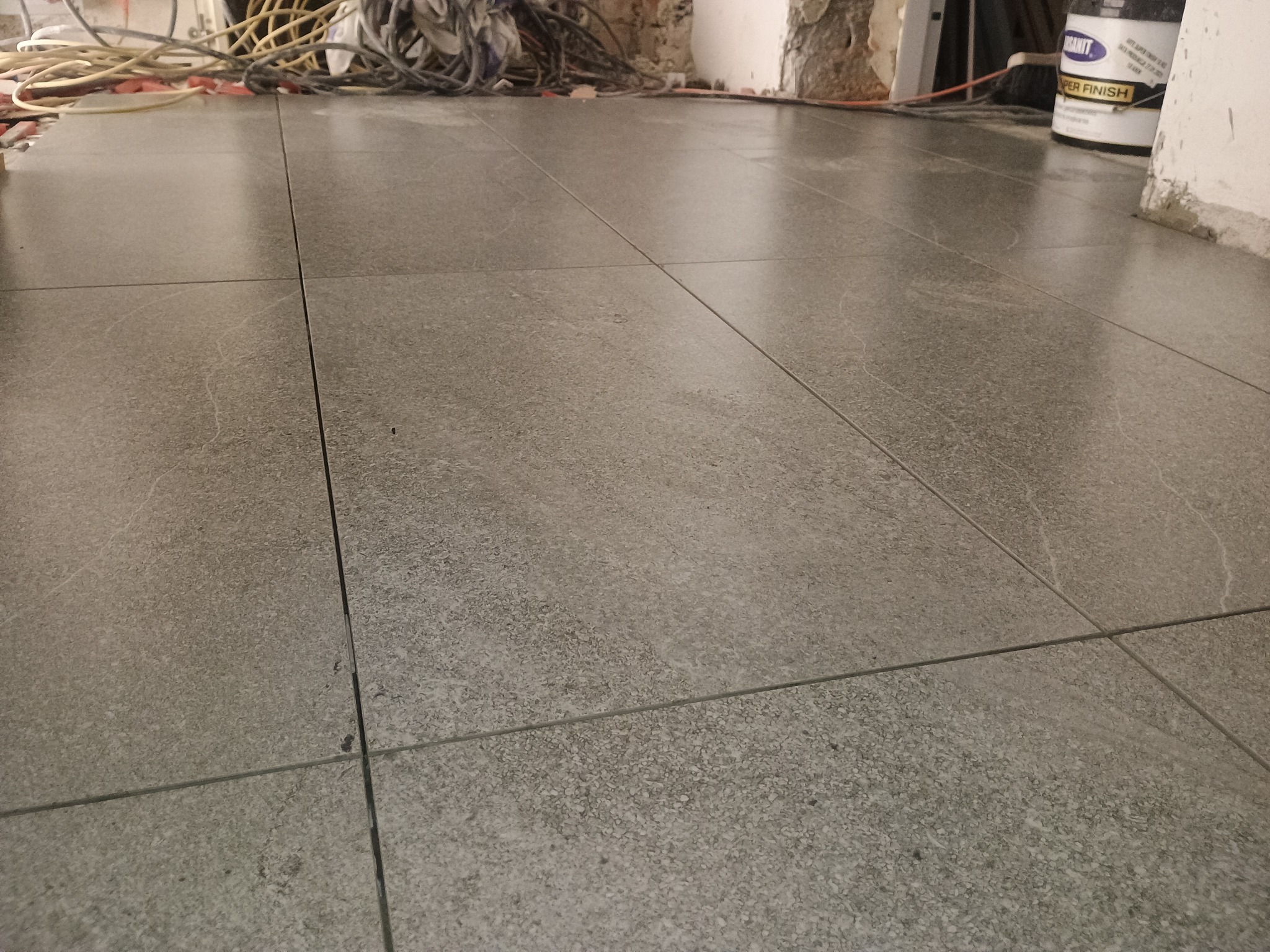 Ułożone, szare płytki podłogowe z widocznymi fugami; w tle kable, ściany w trakcie wykończenia i wiadro z napisem 'Super Finish'.