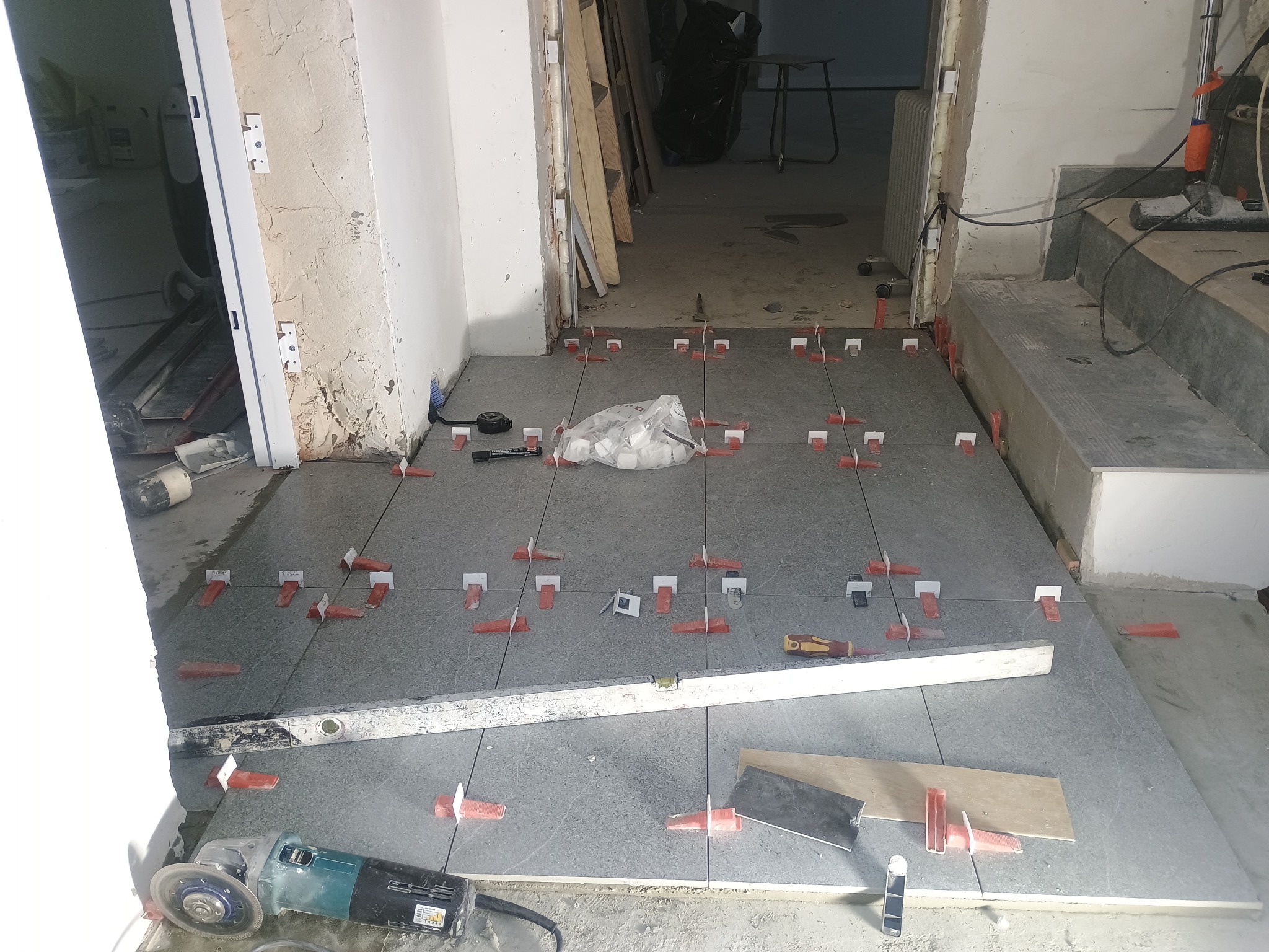 Układanie szarych płytek podłogowych, użycie poziomicy i klipsów dystansowych. Widoczna szlifierka kątowa i torba z klinami do płytek. Wnętrze w trakcie prac wykończeniowych.