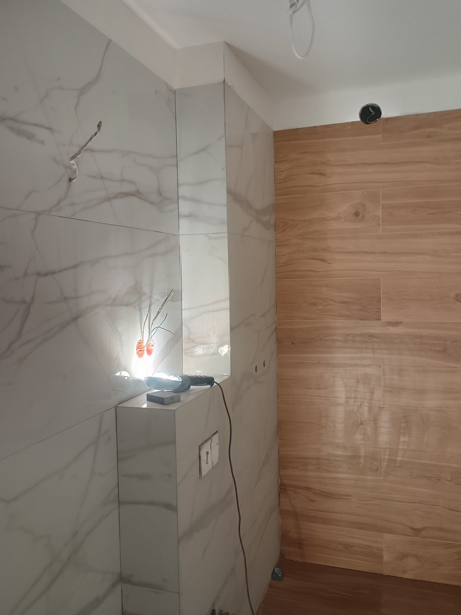 Układanie płytek w łazience: marmurowe i drewnopodobne. Widoczne narzędzia i puszki elektryczne. Detal wykończenia, etap prac glazurniczych.