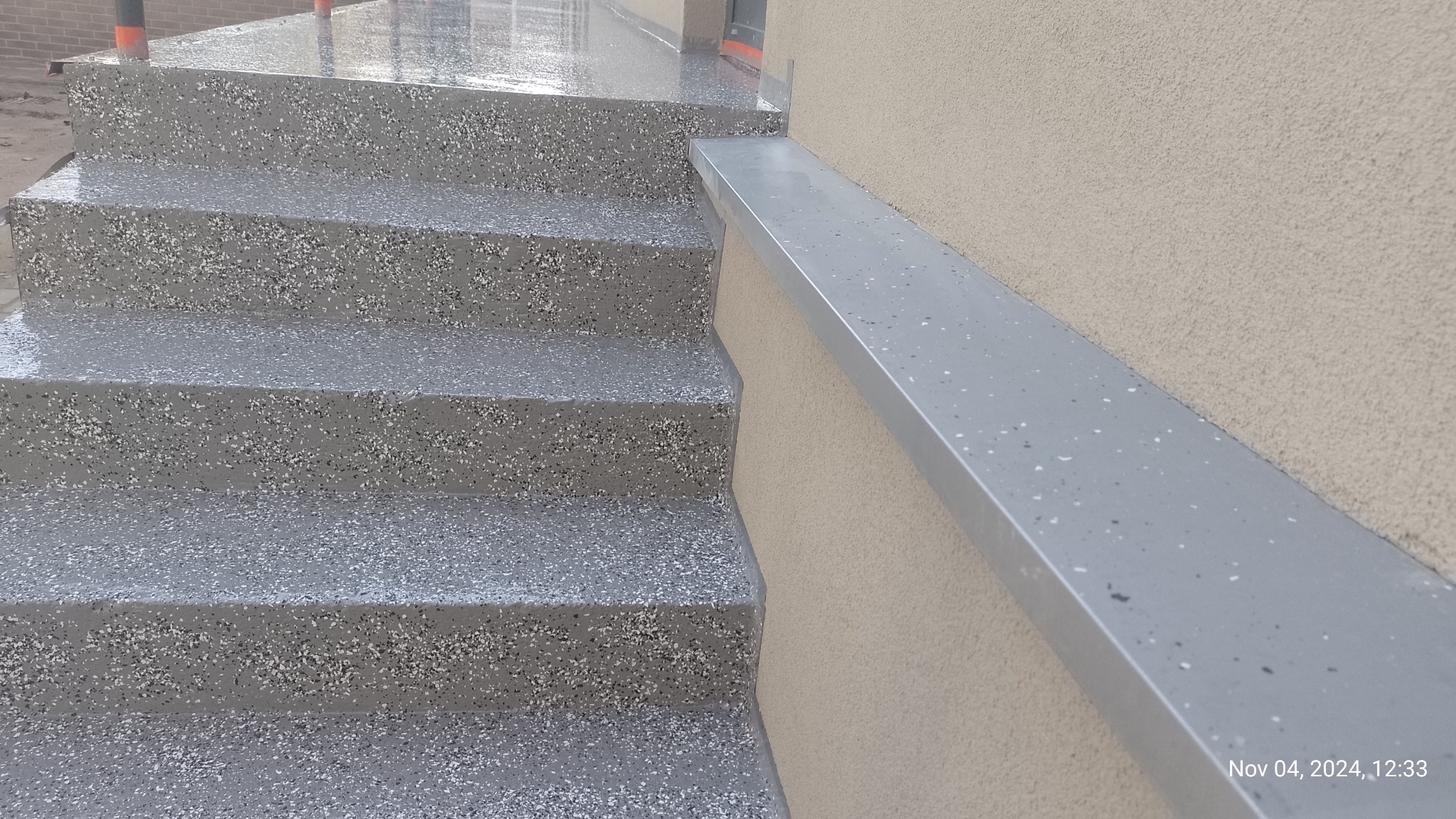 Schody zewnętrzne wykończone szarą, żywiczną posadzką z drobnym, kolorowym kruszywem. Widoczny fragment elewacji z poziomym elementem wykończeniowym.