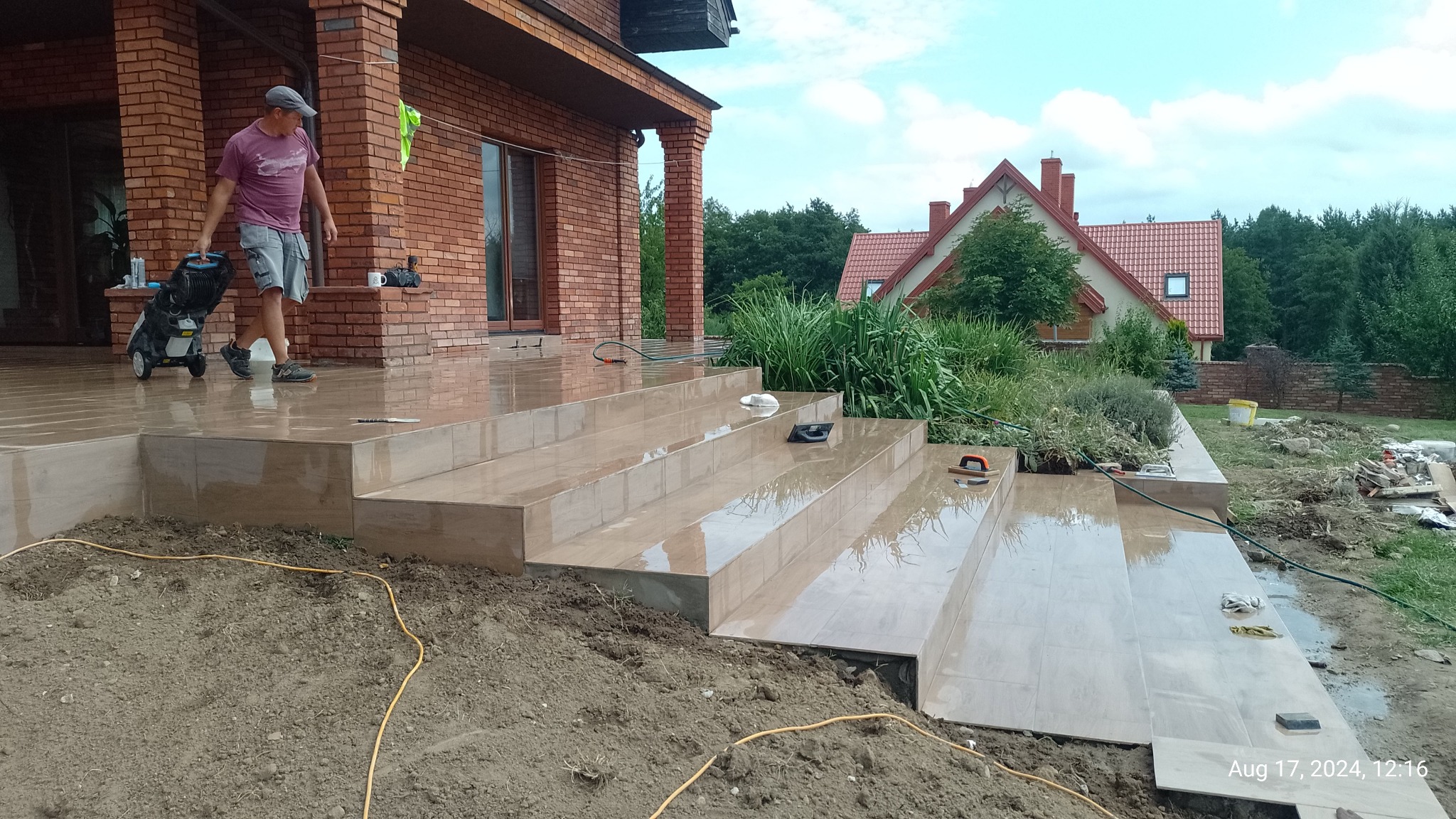 Wyłożone jasnymi płytkami schody zewnętrzne domu jednorodzinnego, częściowo w ziemi. Mężczyzna ciągnie myjkę ciśnieniową. Widoczne narzędzia glazurnicze.