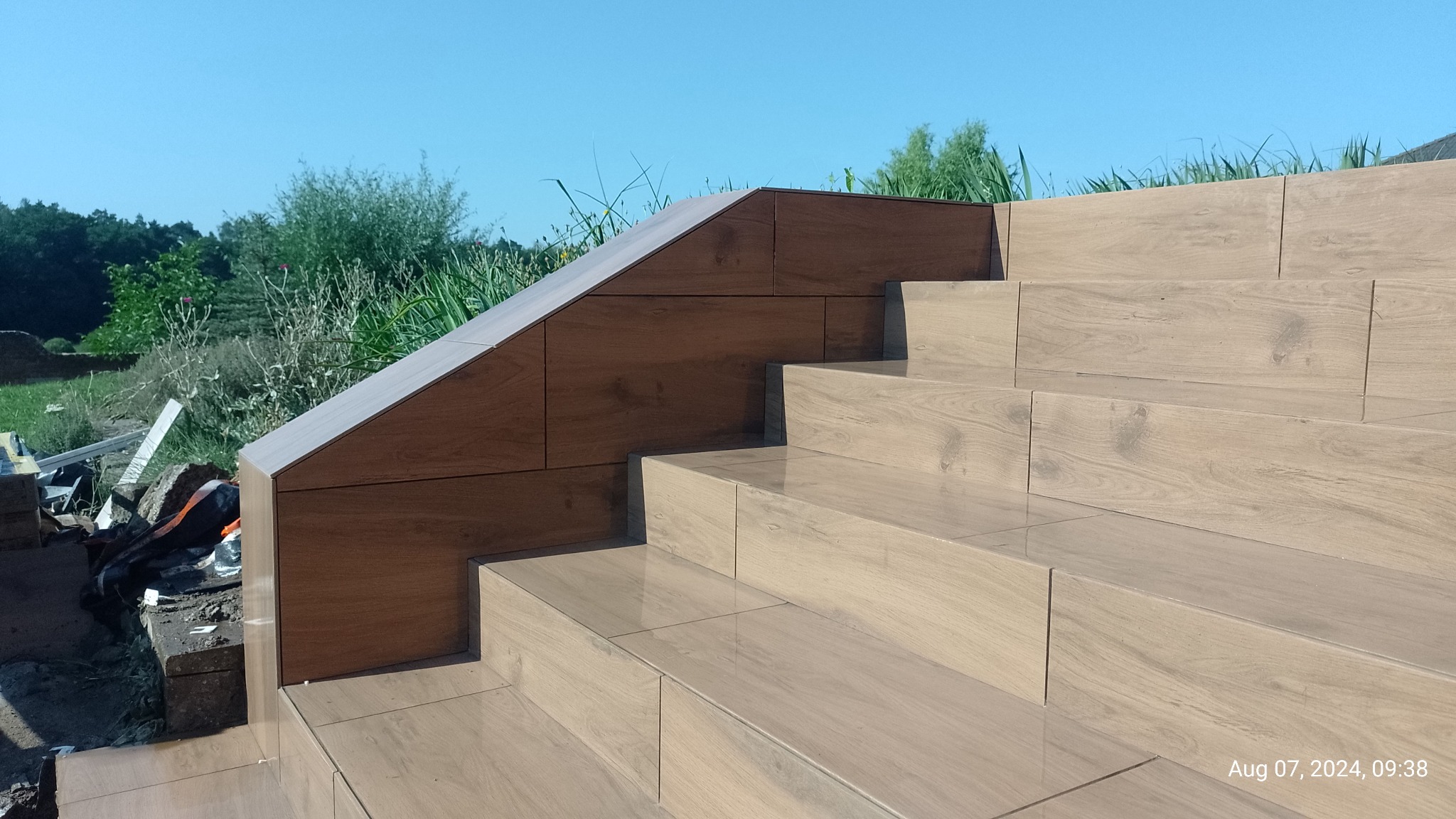 Wykończone płytkami schody zewnętrzne z drewnopodobnym wzorem, prowadzące do ogrodu pod błękitnym niebem. Widoczne detale łączeń płytek i krawędzie schodów.