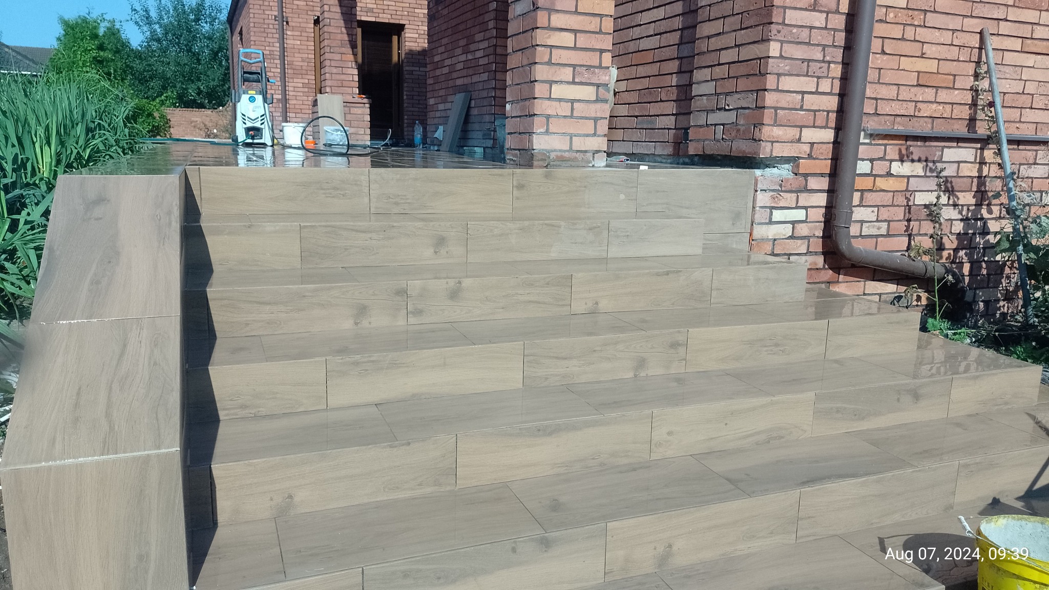 Kafelkowane schody wejściowe do domu, wykończone w odcieniach beżu z delikatnym wzorem drewna. W tle ceglana elewacja i myjka ciśnieniowa.