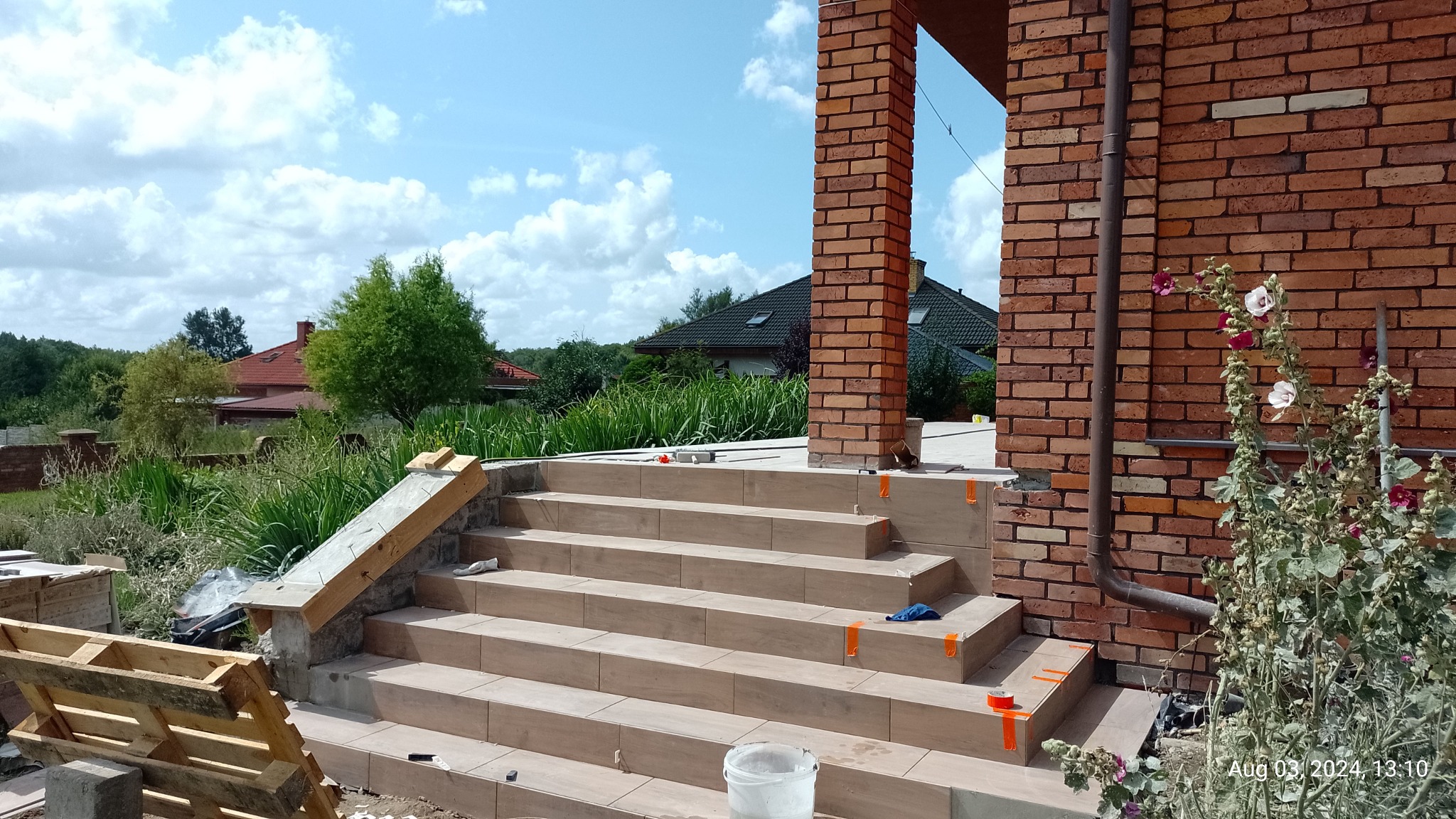 Wyłożone płytkami schody zewnętrzne z ceglaną kolumną w tle, widoczne narzędzia i fragment elewacji. Prace glazurnicze w toku, z detalami wykończeniowymi.