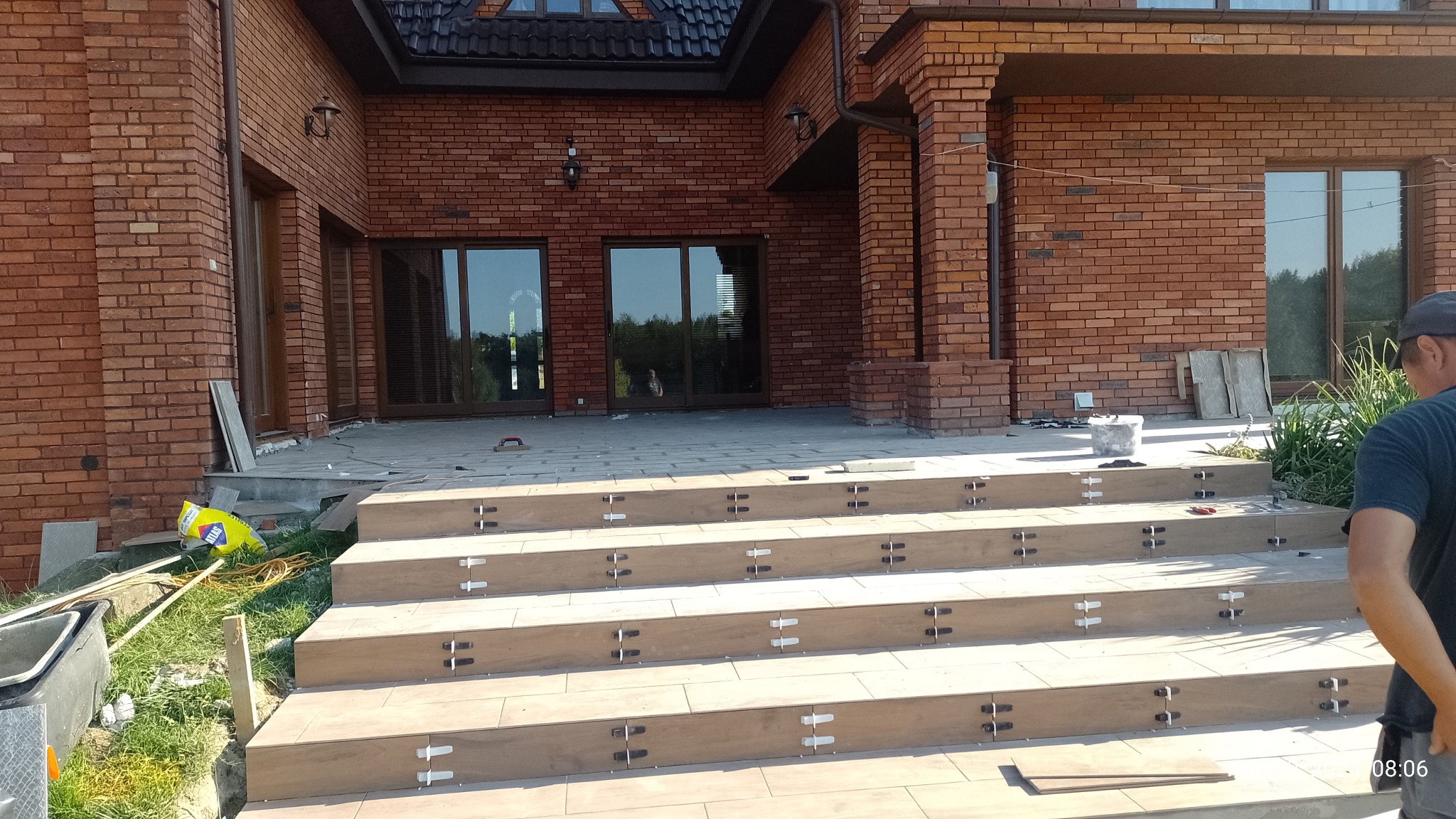 Kaskadowe schody z płytek imitujących drewno, z widocznymi krzyżykami dystansowymi, prowadzące do wejścia domu z czerwonej cegły, w trakcie prac wykończeniowych.