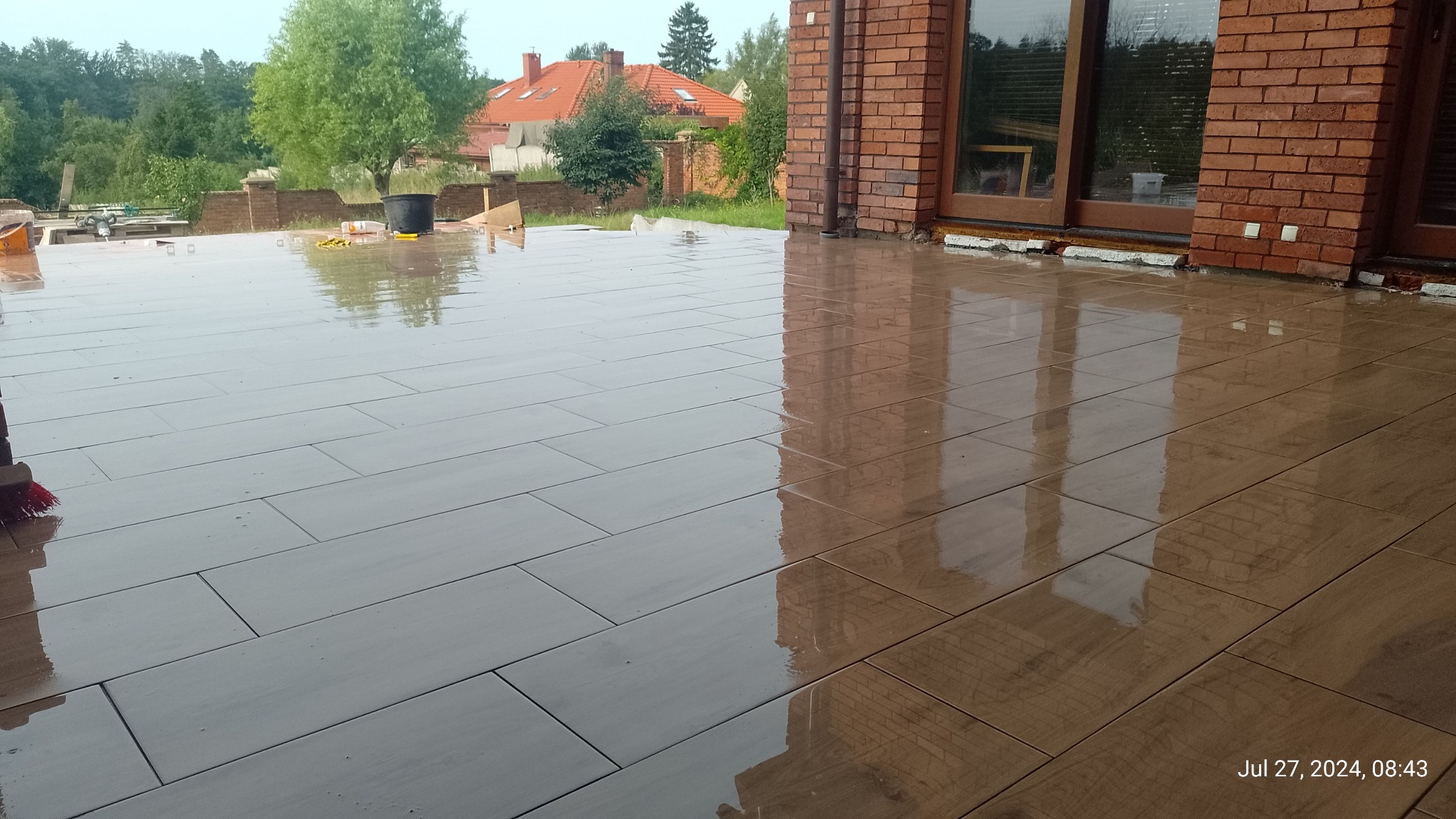 Świeżo ułożona, mokra, prostokątna terakota na tarasie domu z cegły, odbijająca otoczenie. Widoczne narzędzia i wiadro w tle.