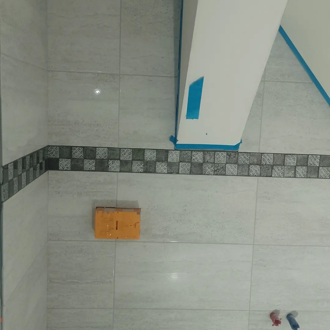 Wykończona ściana w łazience w Słupsku z jasnymi płytkami i dekoracyjnym pasem. Widoczny fragment pionowej konstrukcji zabezpieczonej taśmą malarską.