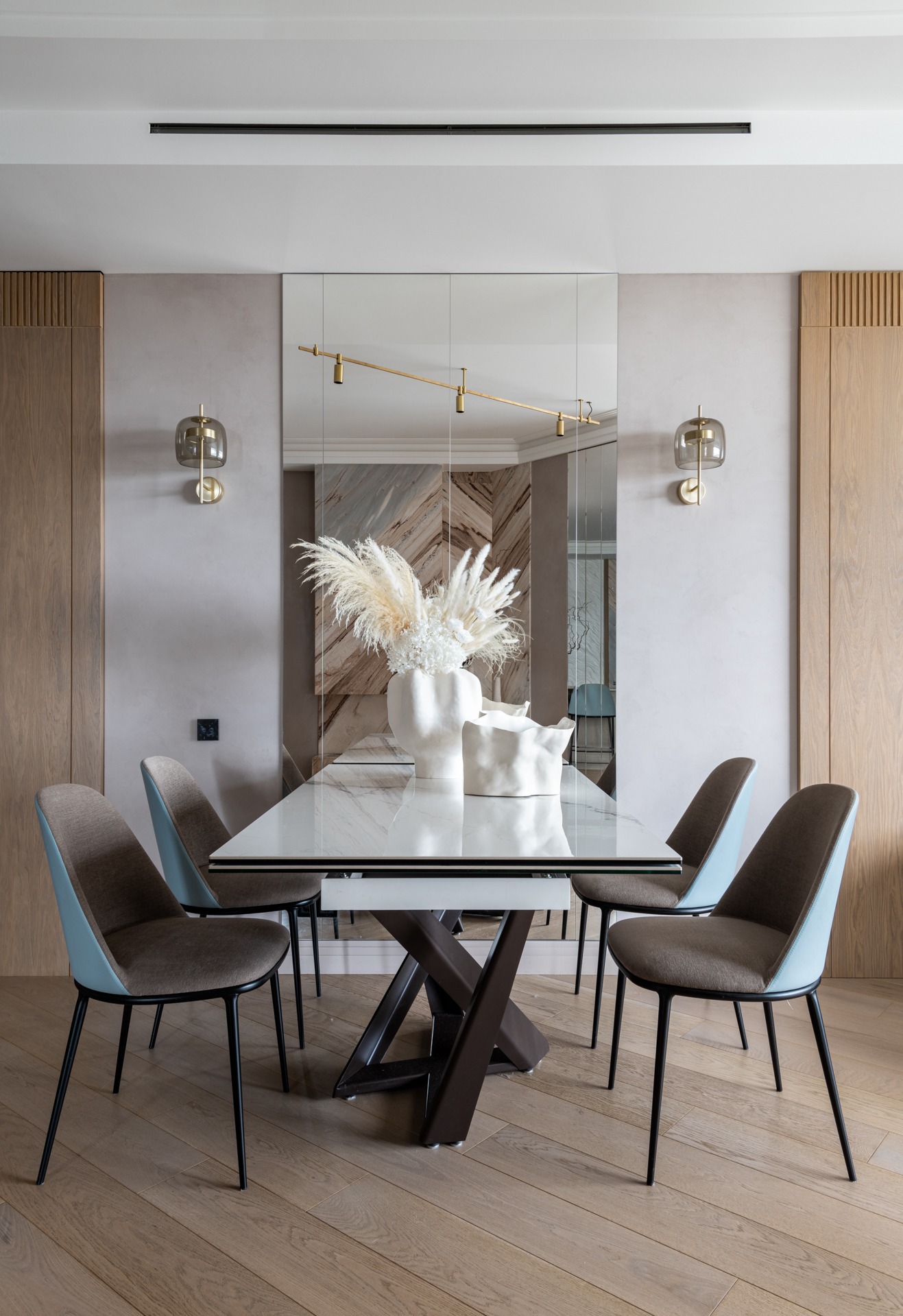 Elegancka jadalnia: stół z białym blatem, designerskie krzesła z błękitnymi oparciami, lustro na ścianie, dekoracyjne lampy ścienne i parkiet w jodełkę.