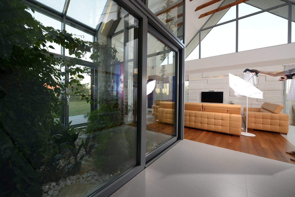 Nowoczesne wnętrze z przeszkloną ścianą, bujną zielenią i beżową sofą. Widoczny sprzęt oświetleniowy i osoba robiąca zdjęcie, co sugeruje sesję fotograficzną.